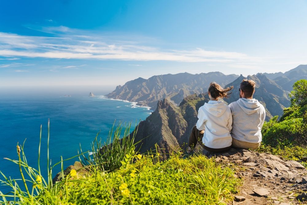 Couple sitting on a cliff, looking at a mountainous coastline and ocean under a bright blue sky.