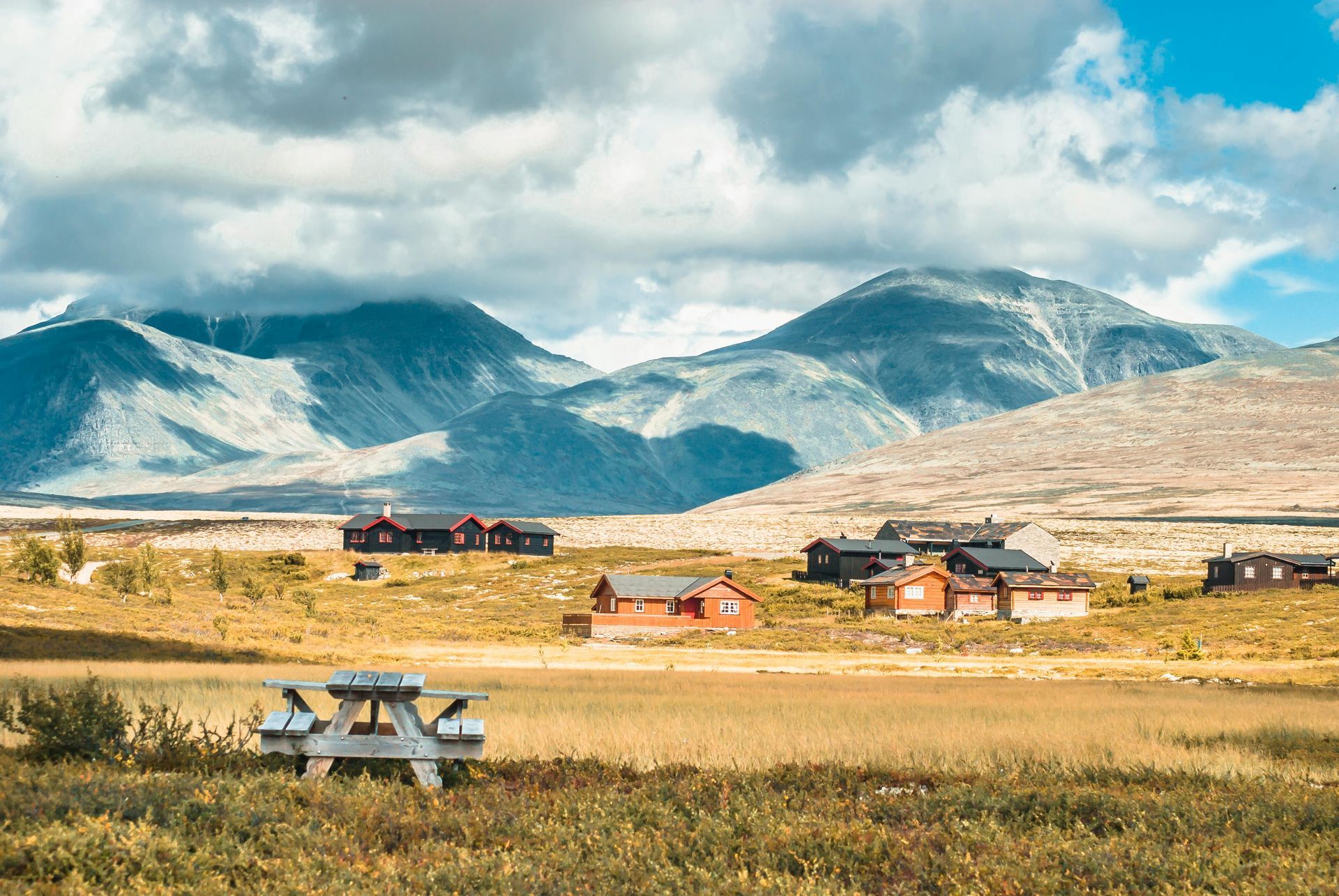 Picnic table in a field with cabins and mountains under a cloudy sky.
