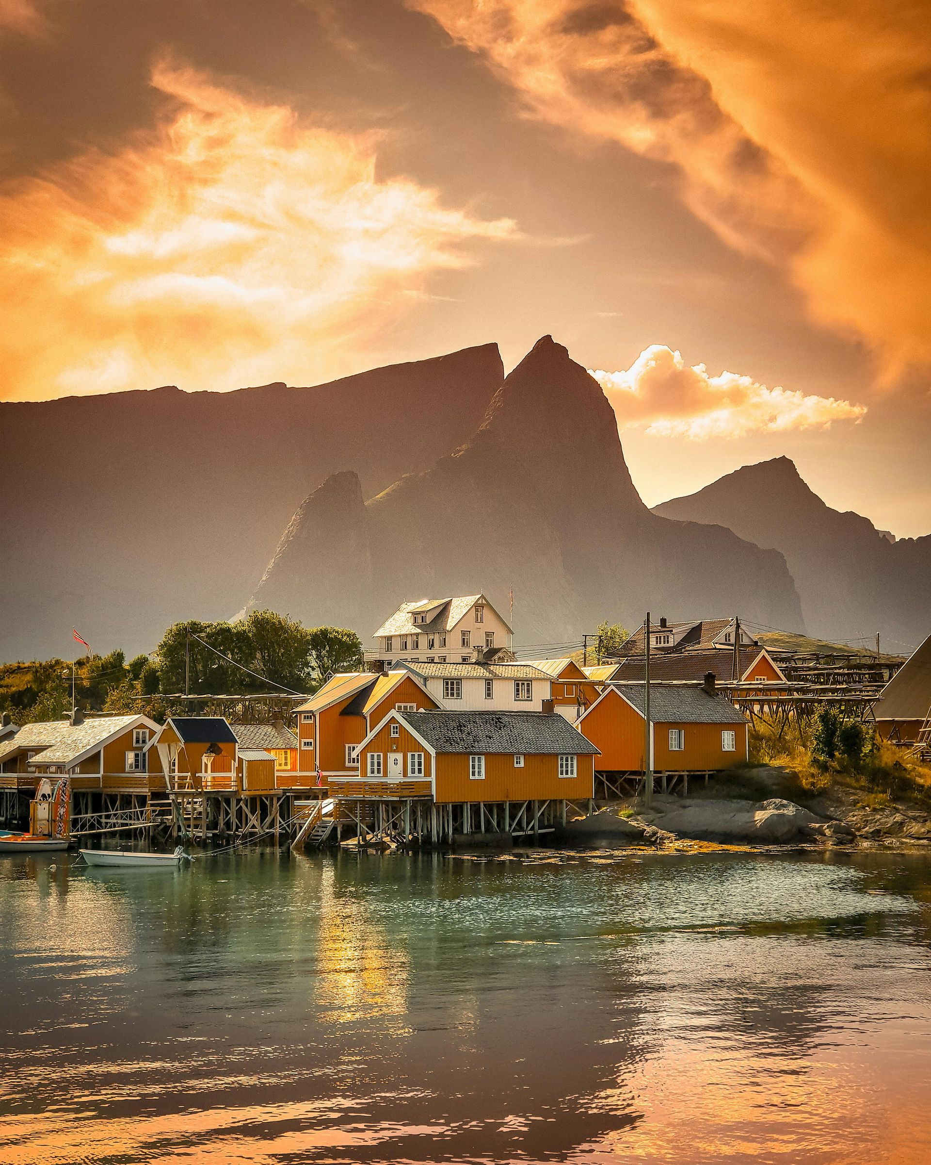 Orange fishing huts on stilts, reflecting in the water, mountains in the background, under an orange sky.