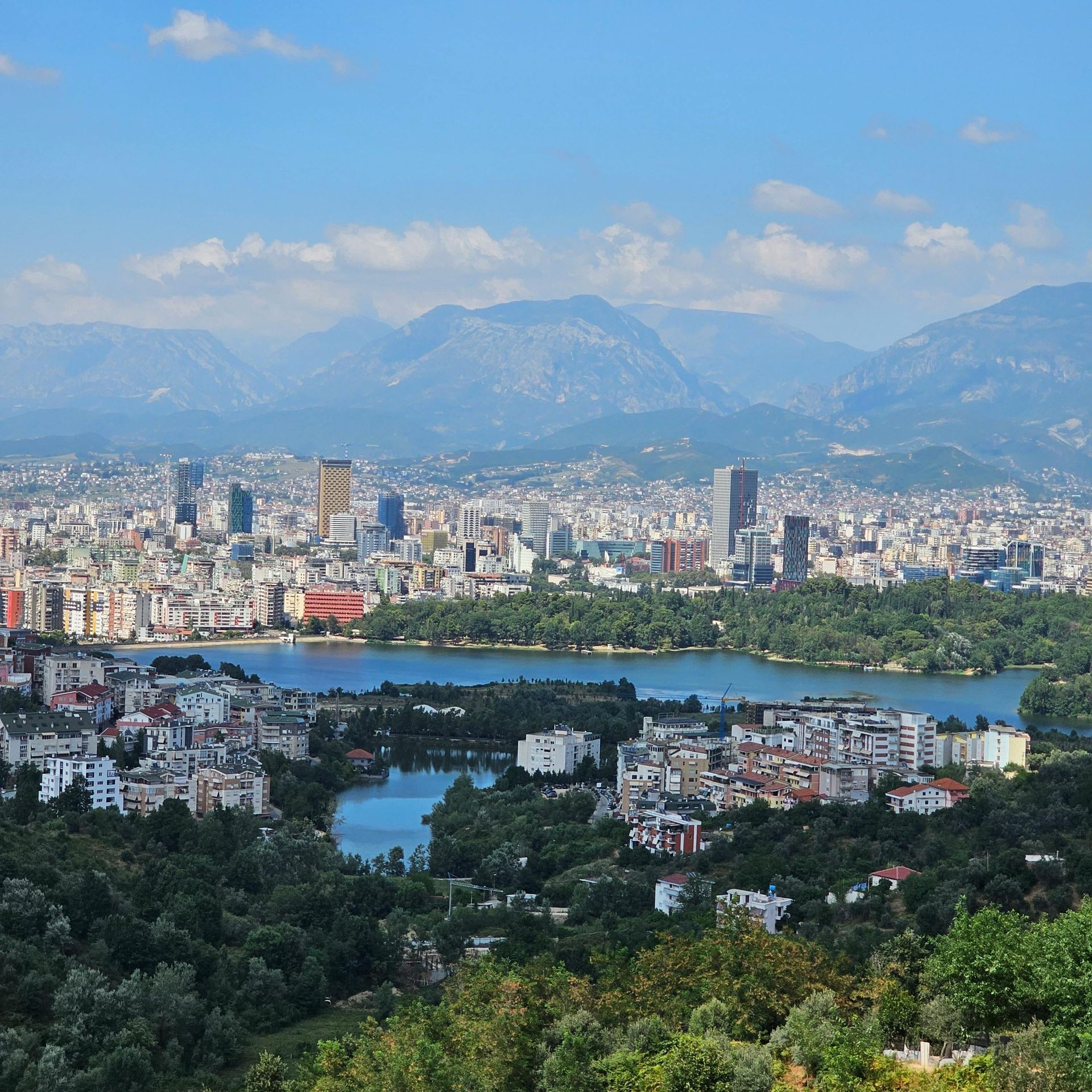 Cityscape overlooking lake, with mountains in the background. Buildings dot the landscape under a blue sky.