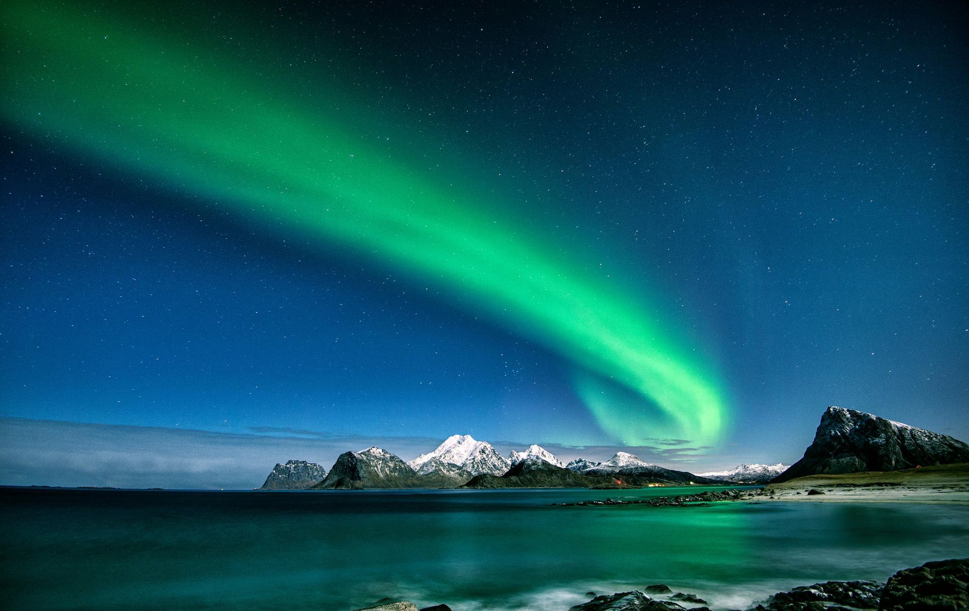 Aurora borealis (Northern Lights) illuminates night sky over snowy mountains and ocean.