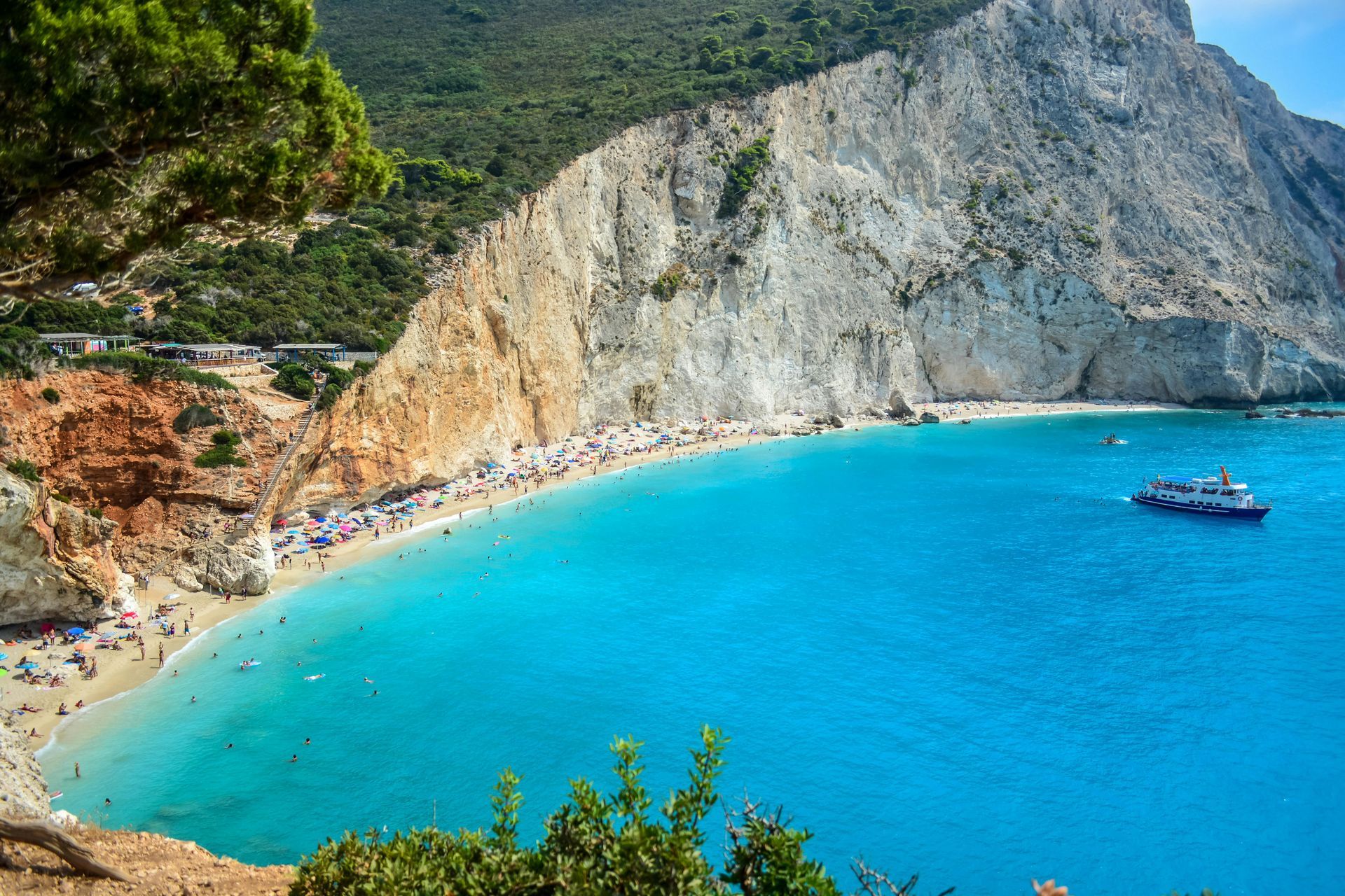 Vibrant blue water meets a sandy beach crowded with people, backed by a white cliff and green hills. A boat floats offshore.