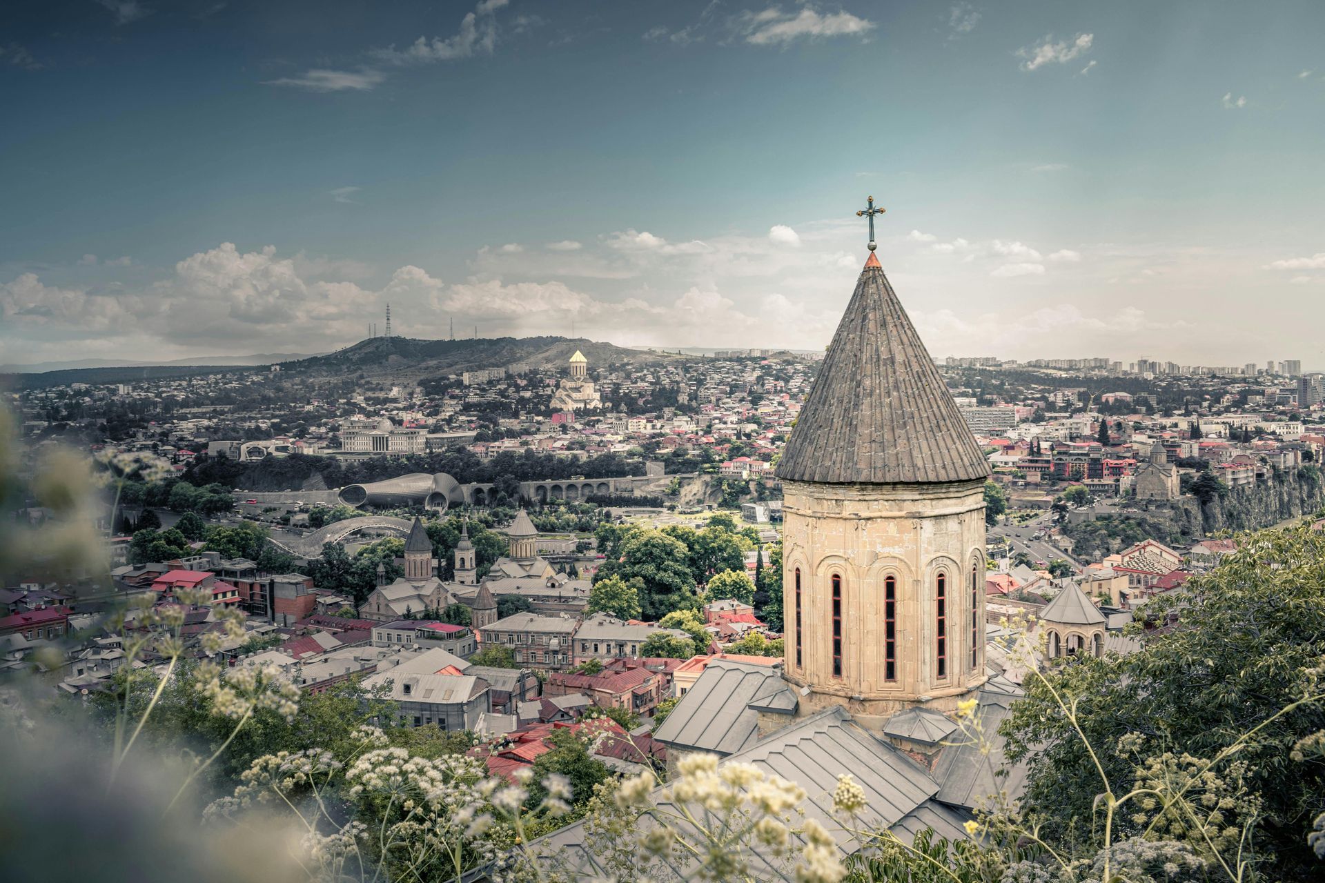 Cityscape with church tower in foreground, buildings and hills in background, under a blue sky.