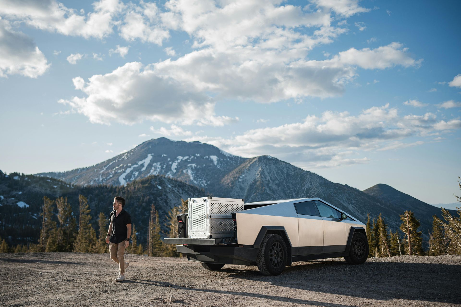A person standing near a white Tesla Cybertruck with a mountain backdrop under a partly cloudy sky.
