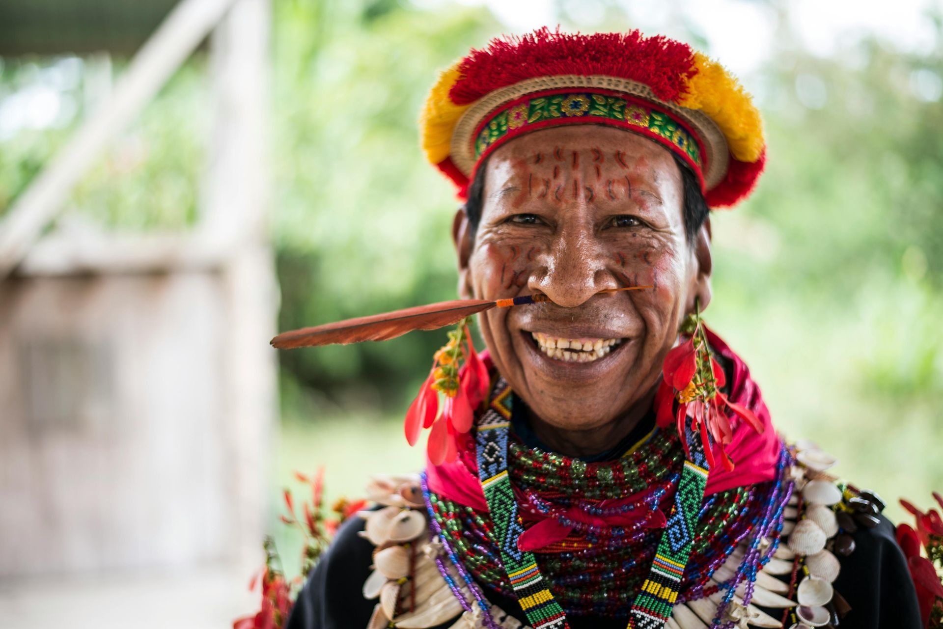 Smiling person wearing a red and yellow headdress, bead necklaces, and a feather in their lip, outdoors.
