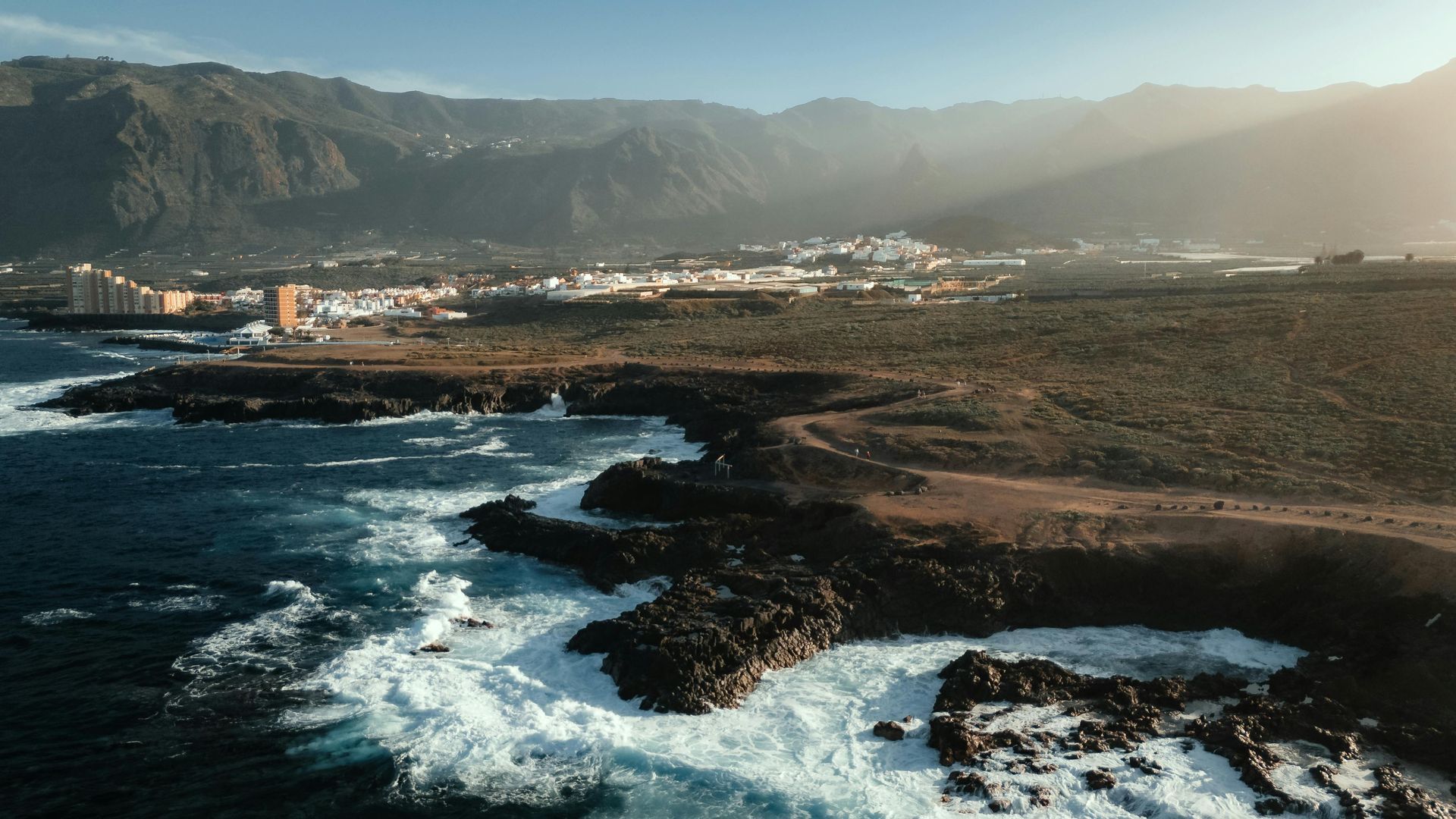 Rocky coastline with ocean waves, town, and mountains under a sunny sky.
