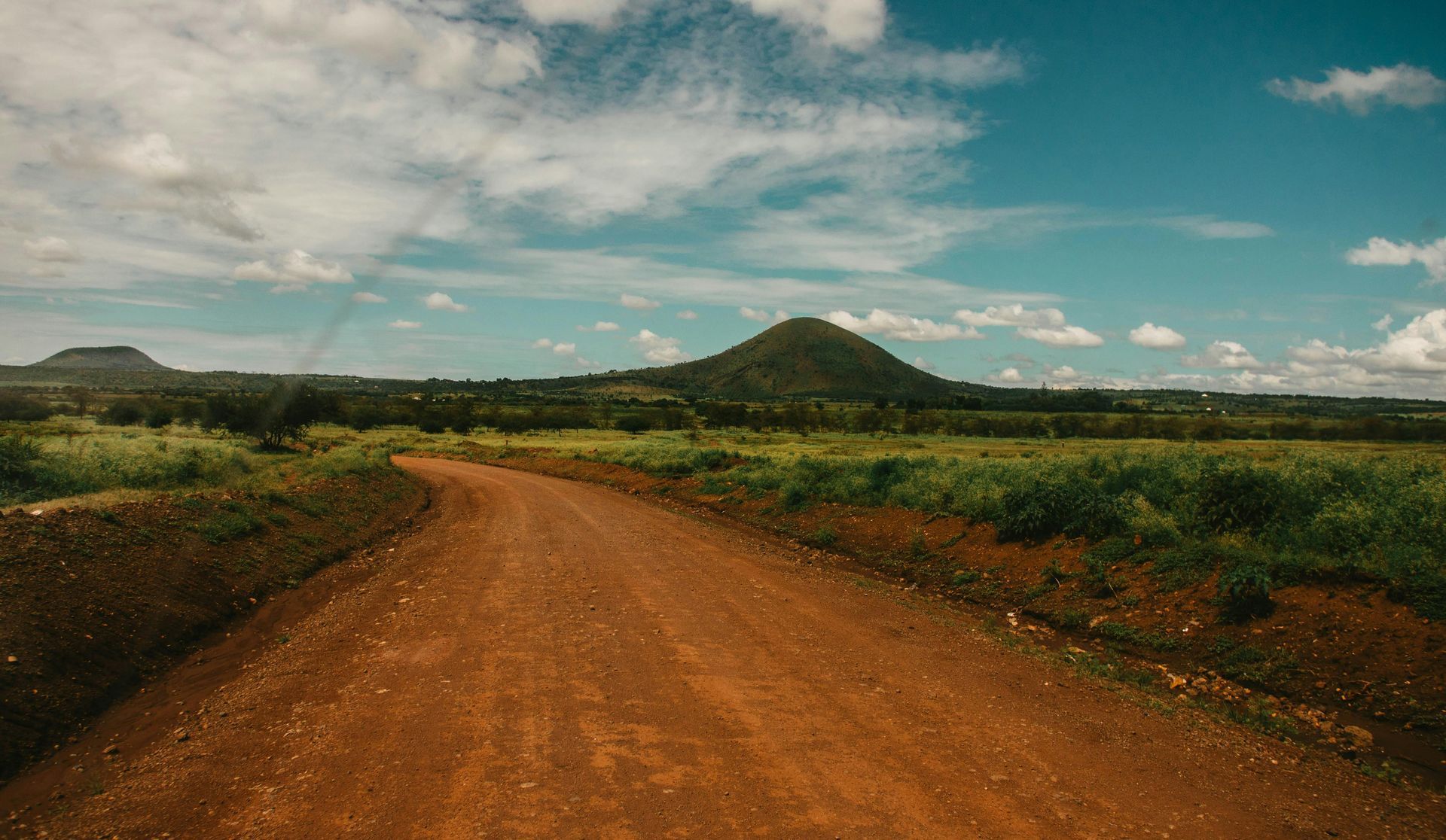 Dirt road leading toward a grassy field and green, rounded hills under a blue sky with scattered clouds.