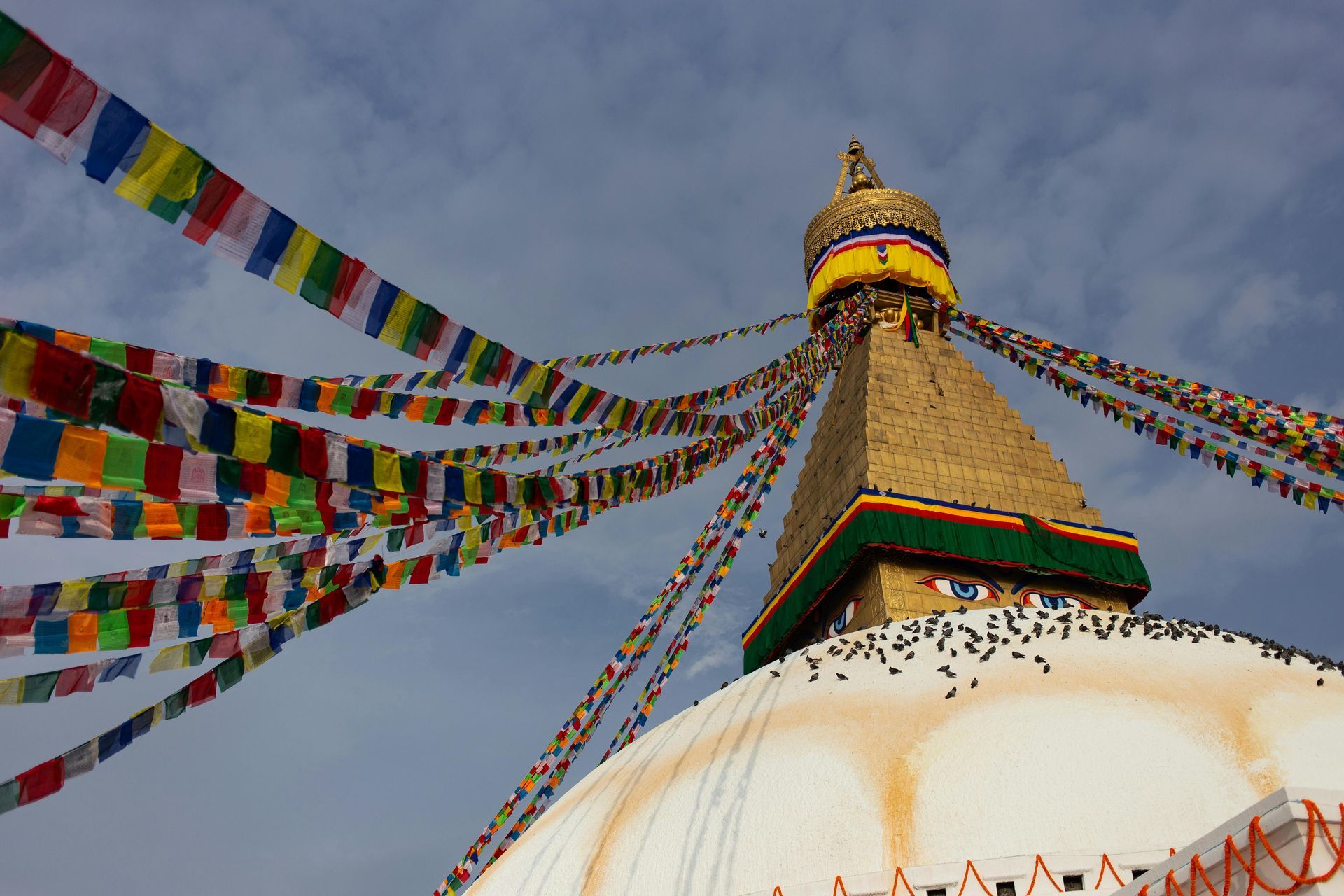 Boudhanath Stupa in Nepal with colorful prayer flags against a cloudy sky.