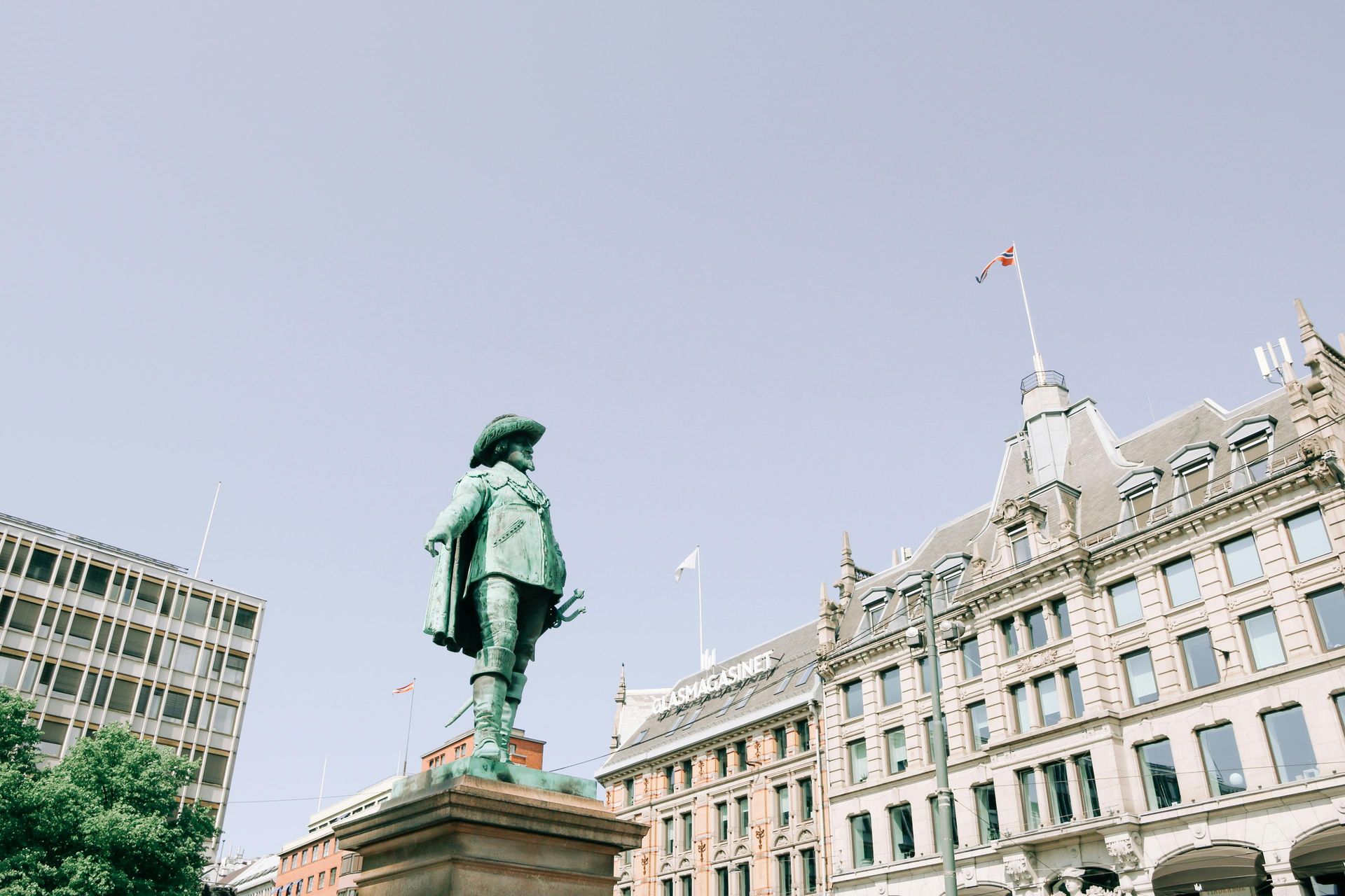 Statue of a man in hat and coat in front of ornate buildings. Sunny day with blue sky.