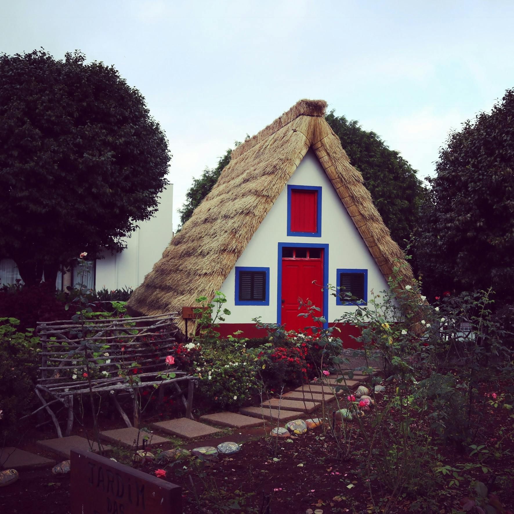 A-frame thatched roof cottage with red door and blue trim, surrounded by flowers.