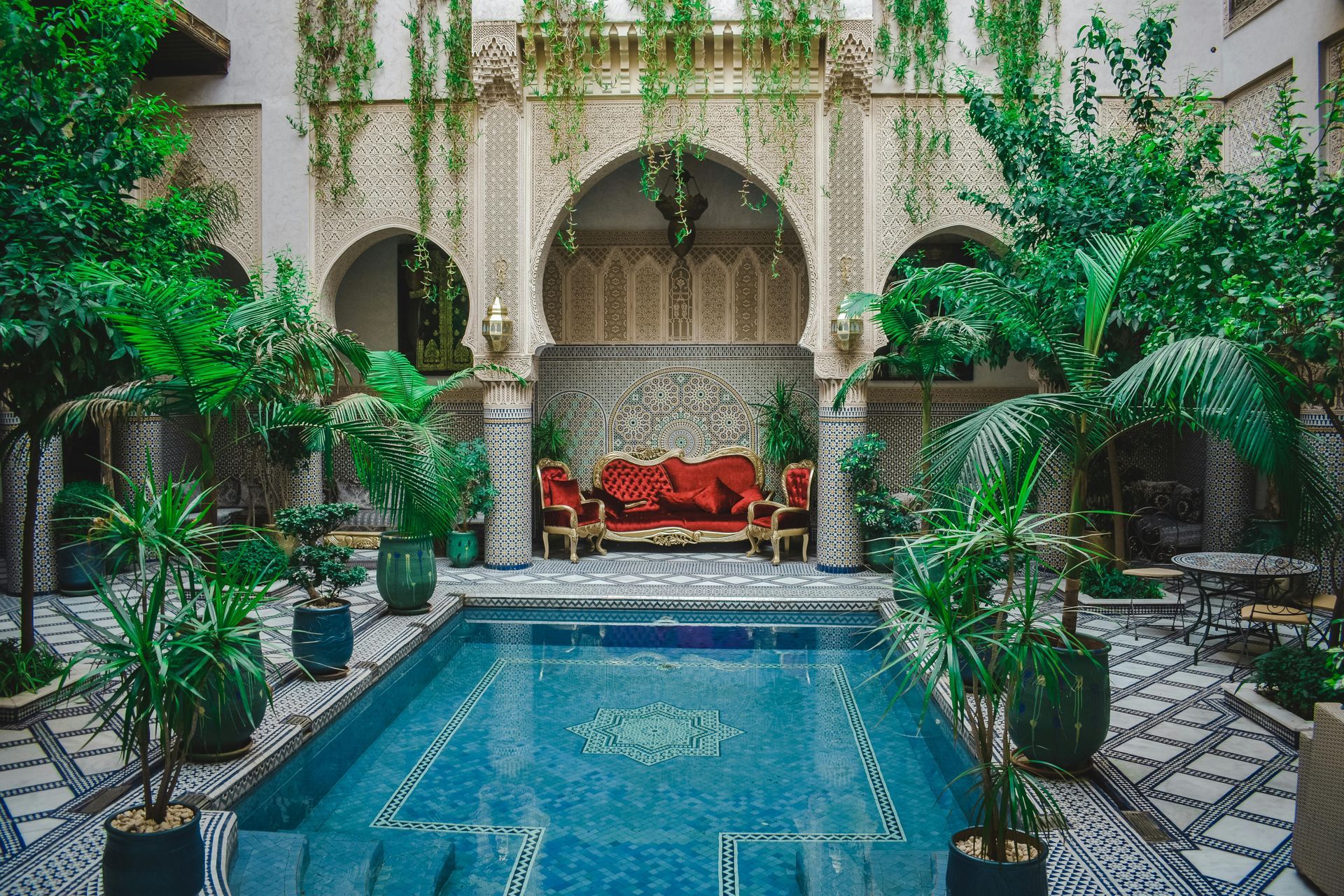 Courtyard with turquoise pool, ornate archways, red sofa, lush greenery, tiled walls.