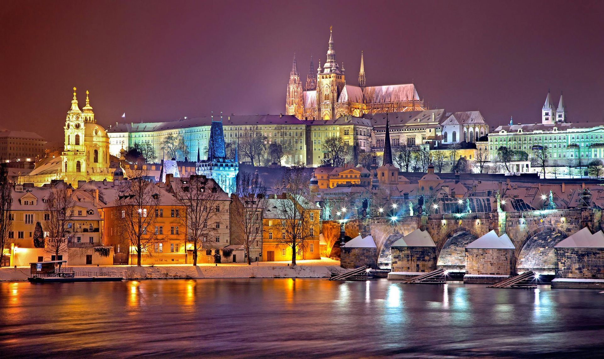 Prague cityscape at night, with illuminated buildings, snowy rooftops, and the Charles Bridge over a dark river.