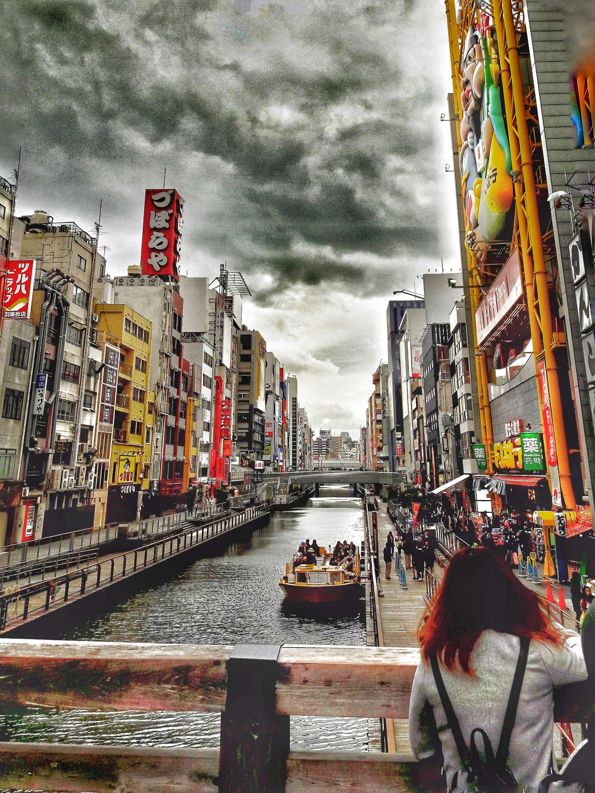 Canal in Osaka with boat, buildings, and overcast sky. Woman with red hair is looking towards the water.