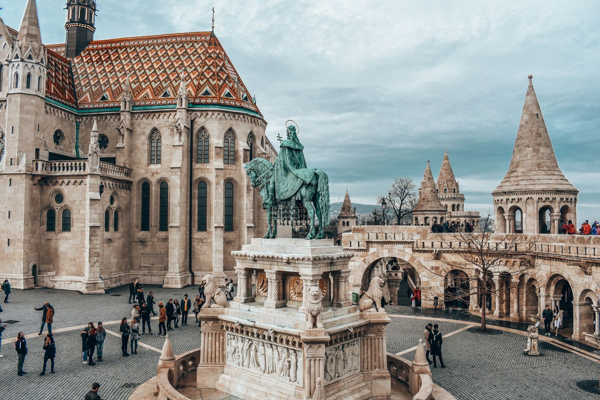 Matthias Church and Fisherman's Bastion in Budapest, Hungary, with statue, tourists, and cloudy sky.