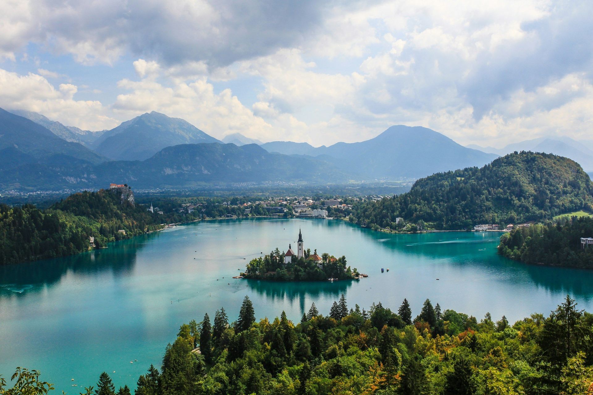 Turquoise Lake Bled, Slovenia, with island church, surrounded by green hills, mountains, and town under cloudy sky.