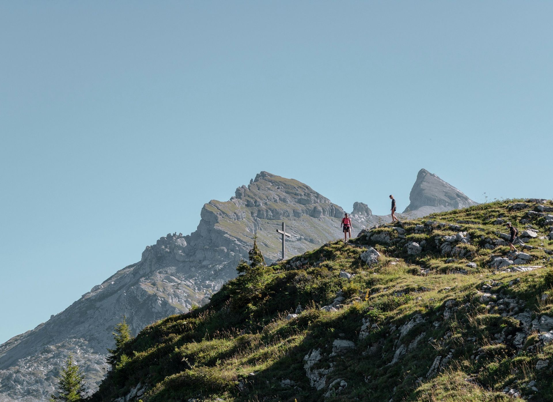 Hikers on a grassy mountain slope with rocky peaks under a clear blue sky.