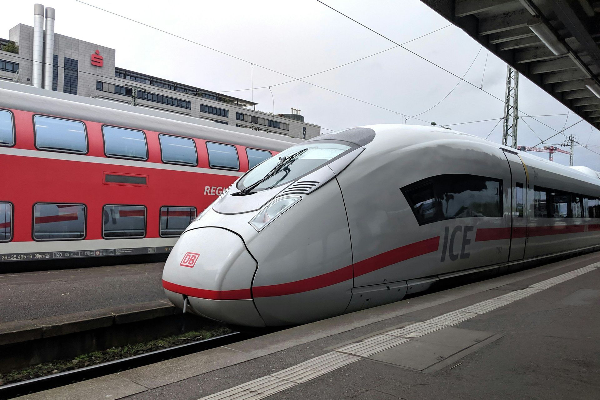 A high-speed ICE train at a station platform, next to a red train car, overcast sky.