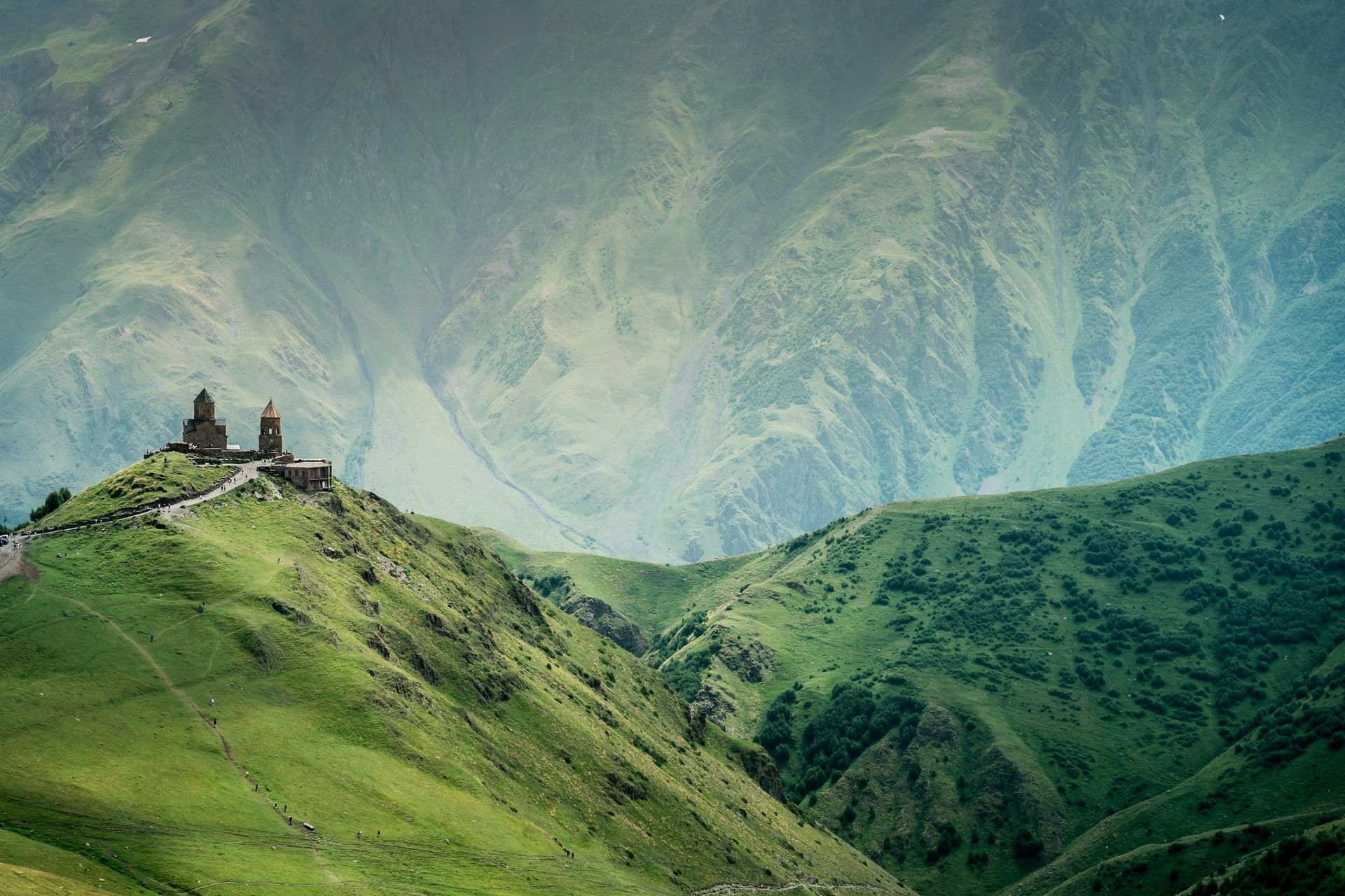 Mountain landscape with a church on a hilltop, green slopes, and misty mountains in the background.