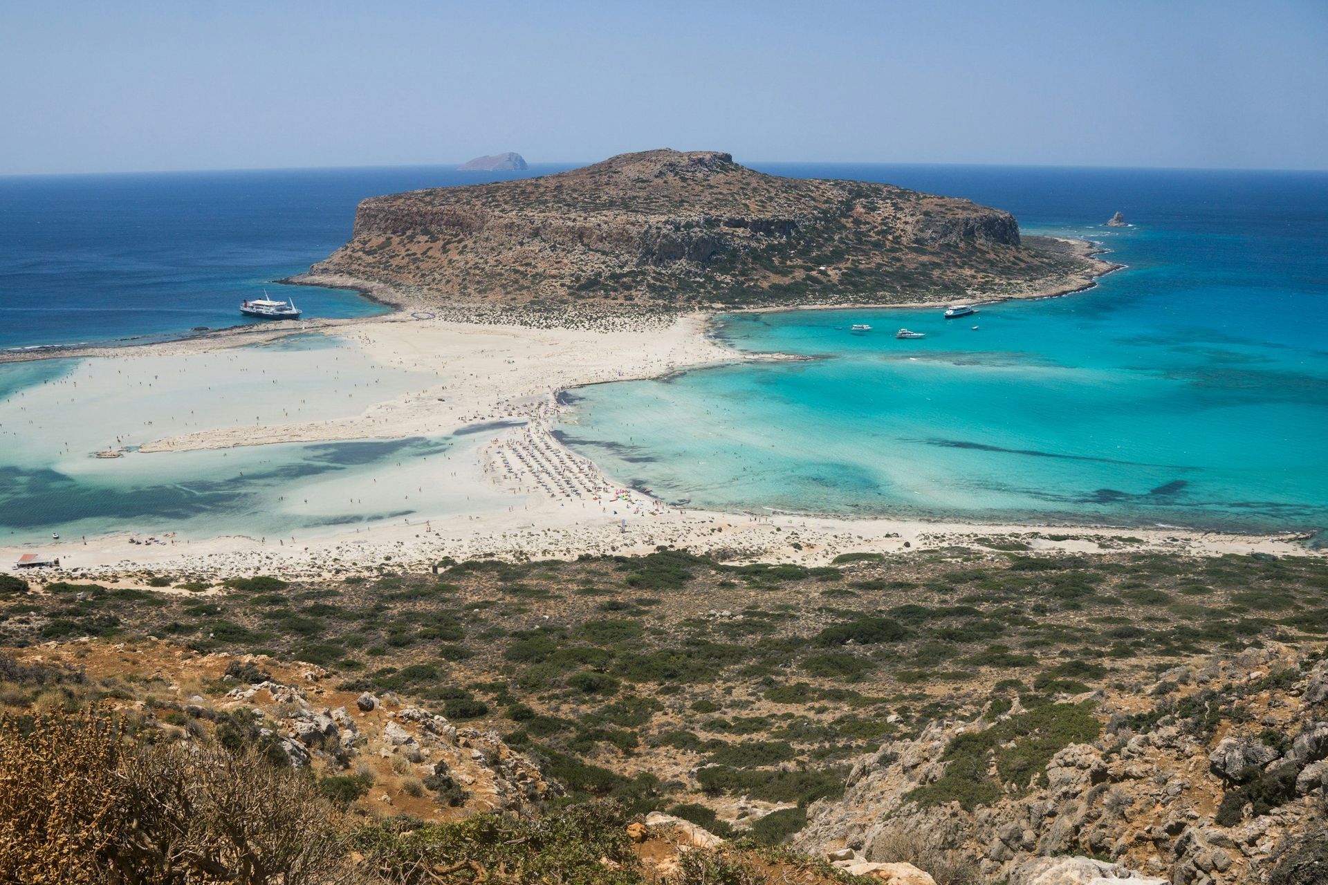 Island with turquoise water, white sandbars, and a rugged, brown islet in the background. People on the beach.