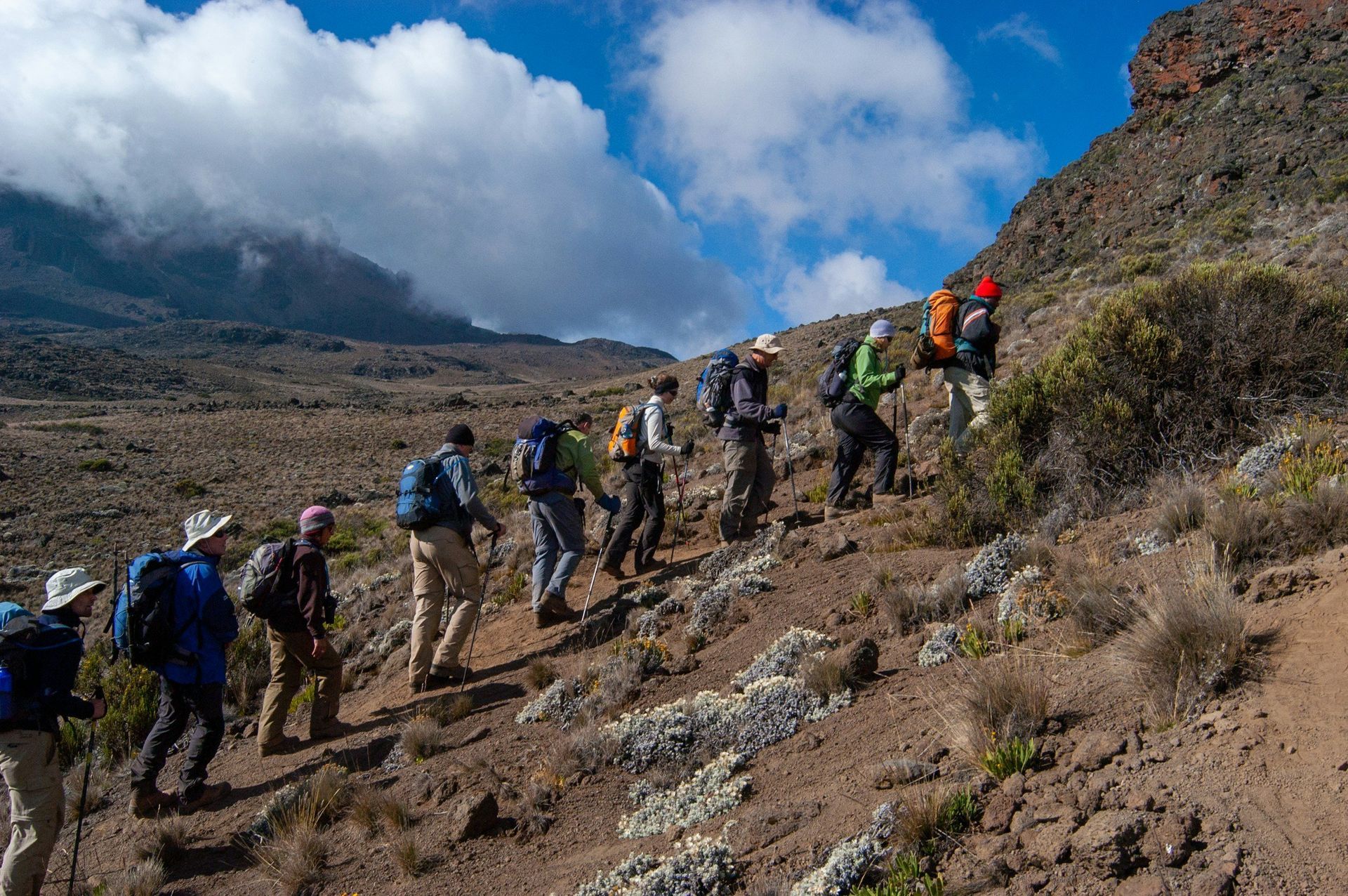Group of hikers ascending a rocky mountain trail under a partly cloudy sky.