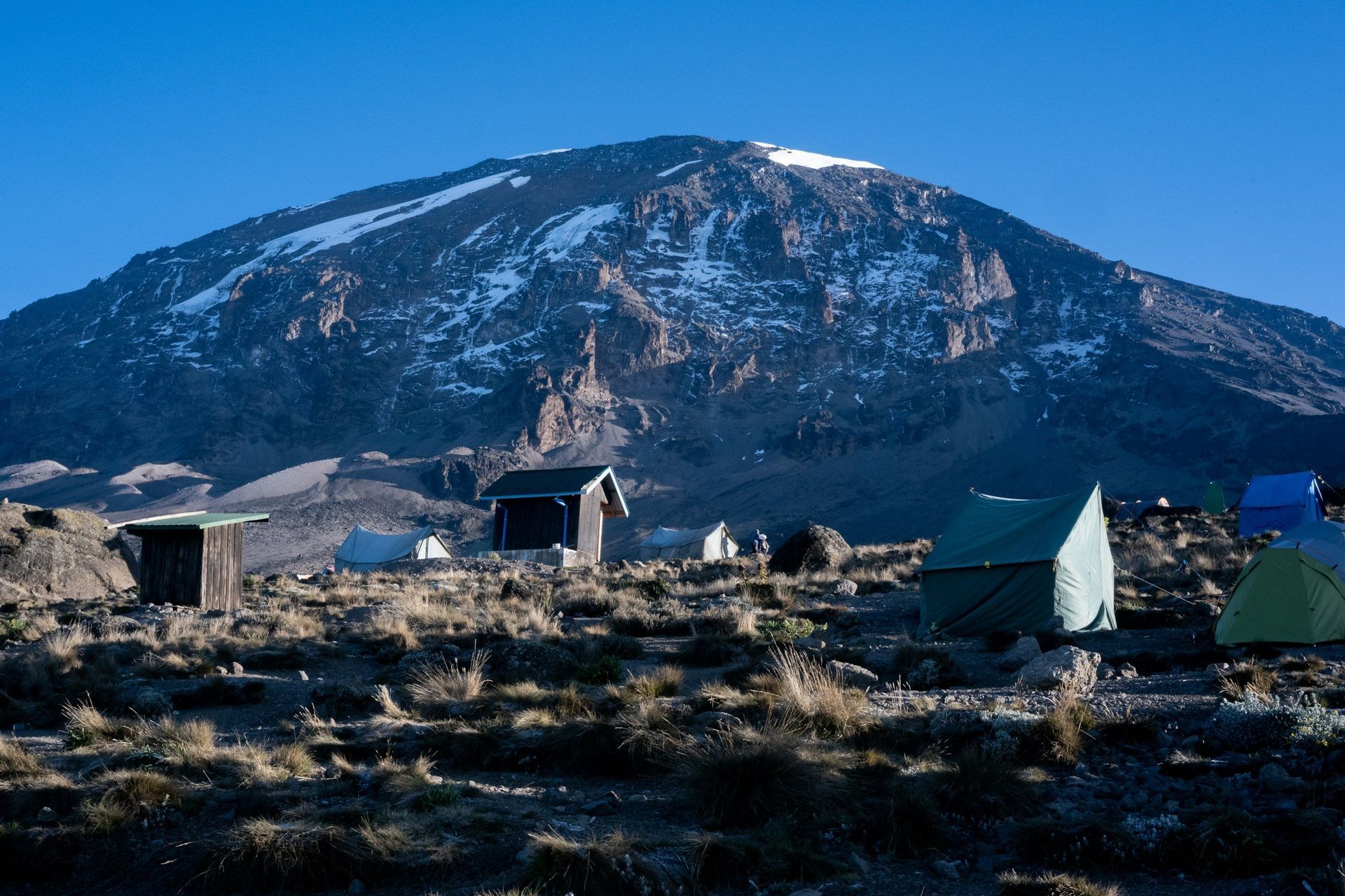 Campsite with tents and outhouses at the base of Mount Kilimanjaro, Tanzania. Snowy peak in the background under a blue sky.