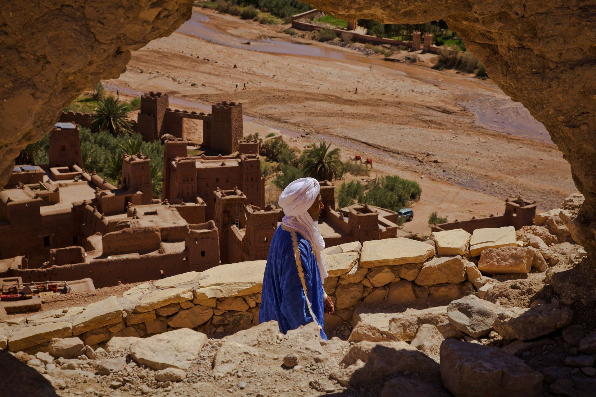 Man in blue robe and turban looks out over ancient mud-brick village in a desert landscape.
