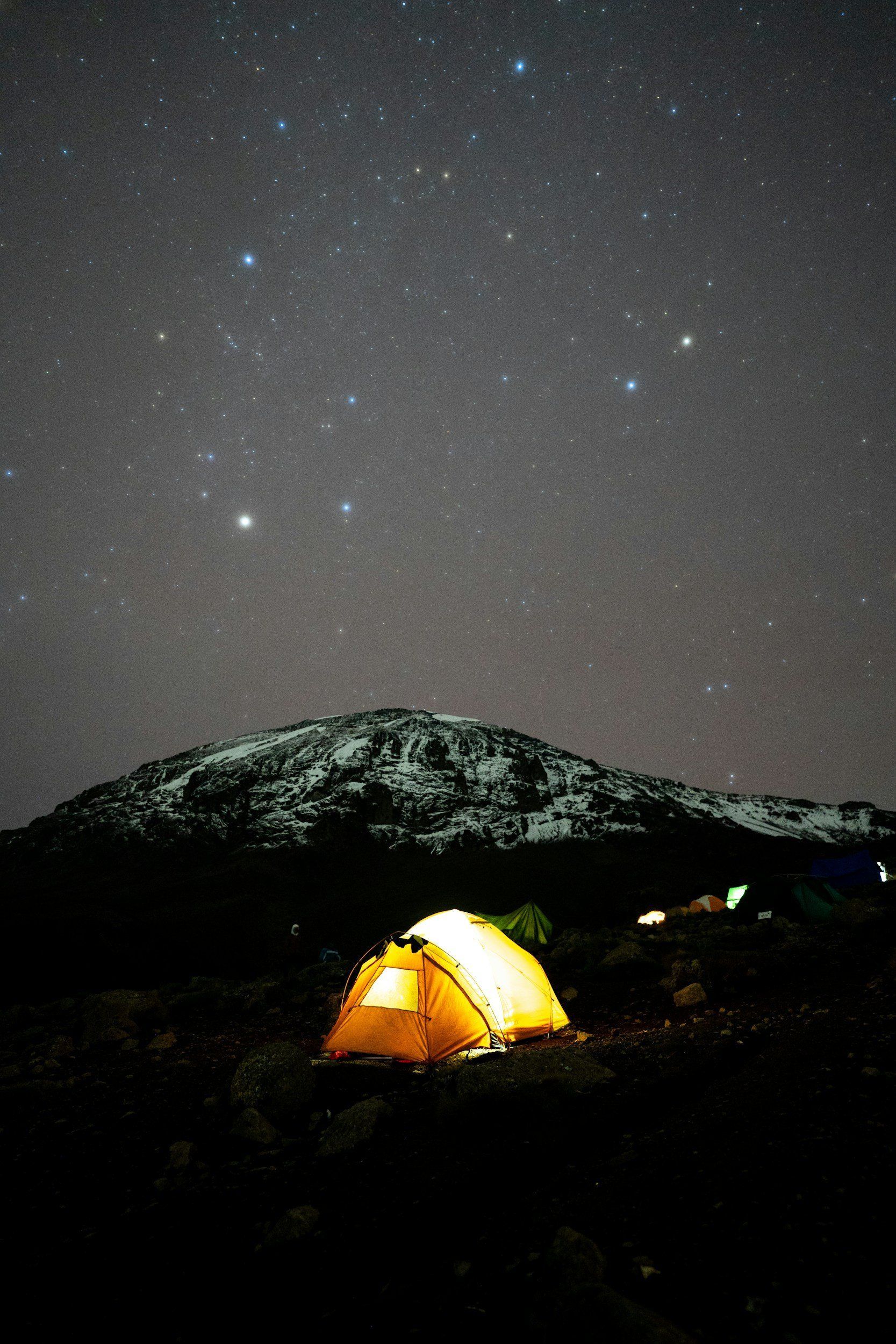A lit tent under a starry night sky, with a snow-dusted mountain in the background.