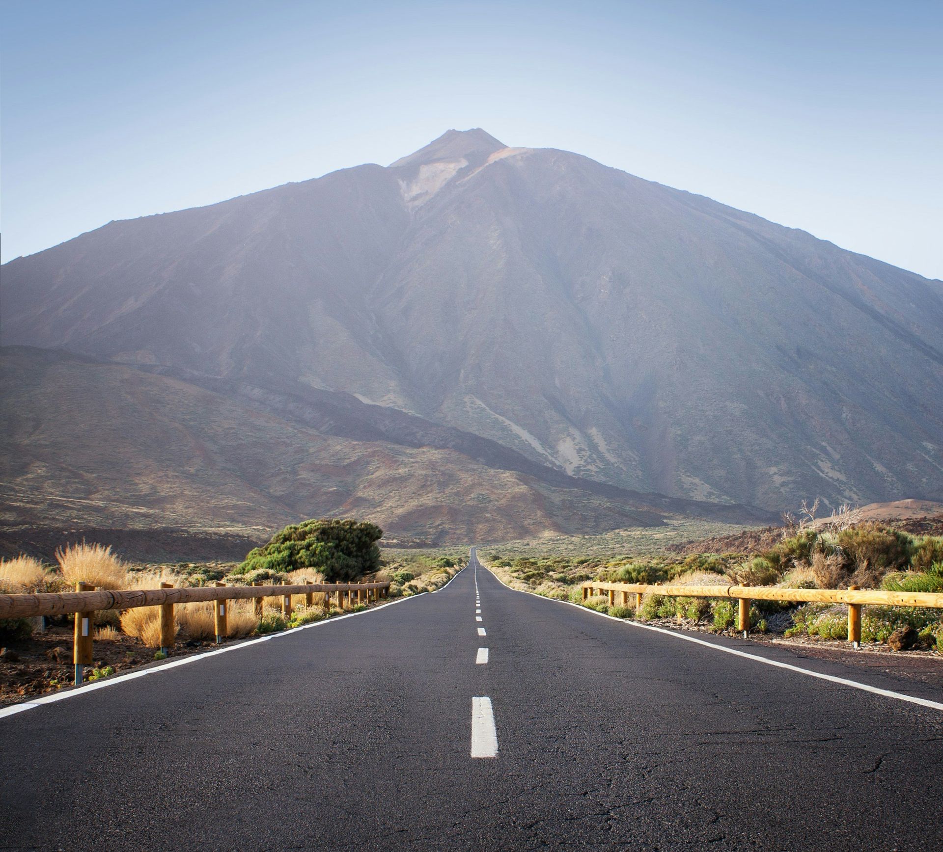 Road stretches towards a large mountain under a clear blue sky.