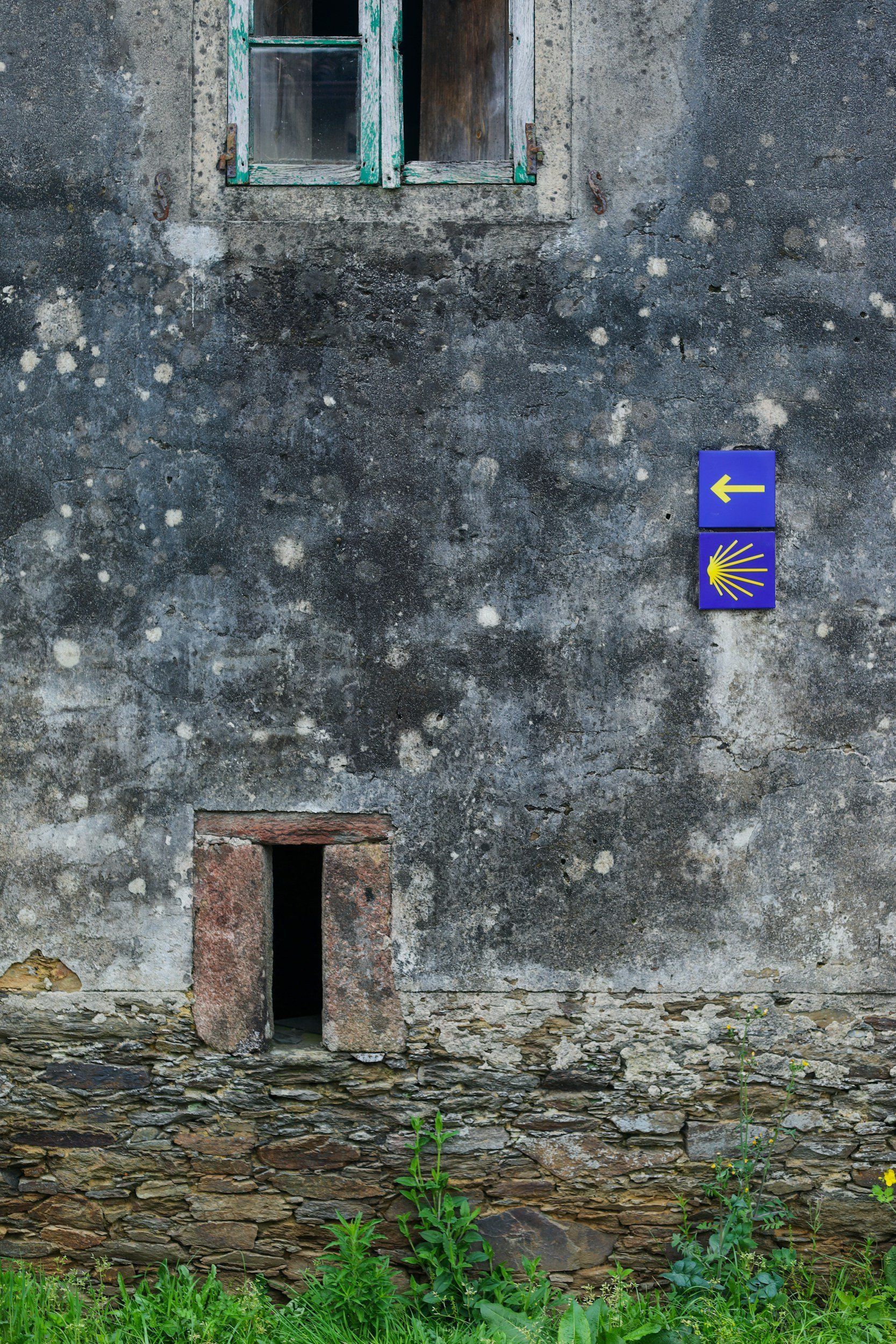 Weathered building facade with window and signs pointing to the left; lower opening and green vegetation below.