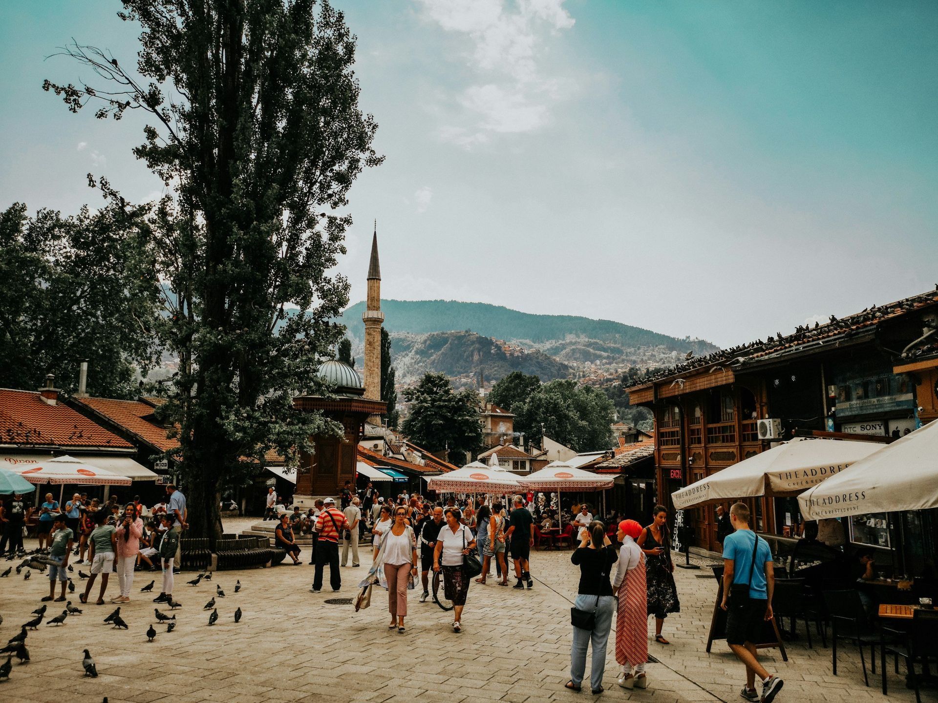 Busy square in Sarajevo, Bosnia, with people walking, a tall mosque minaret, and shops on a sunny day.