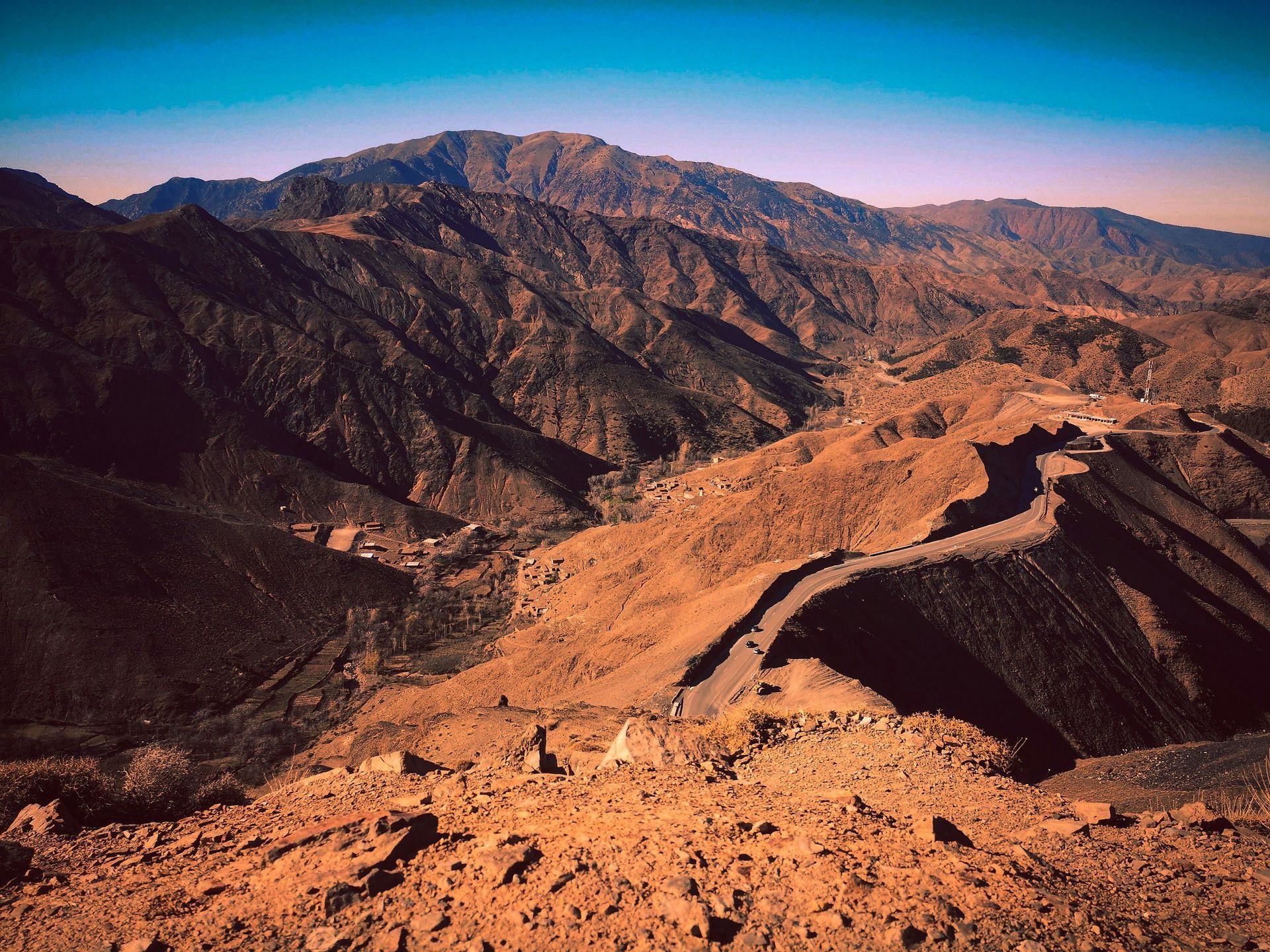 Mountain range with a winding road; earthy tones, blue sky.