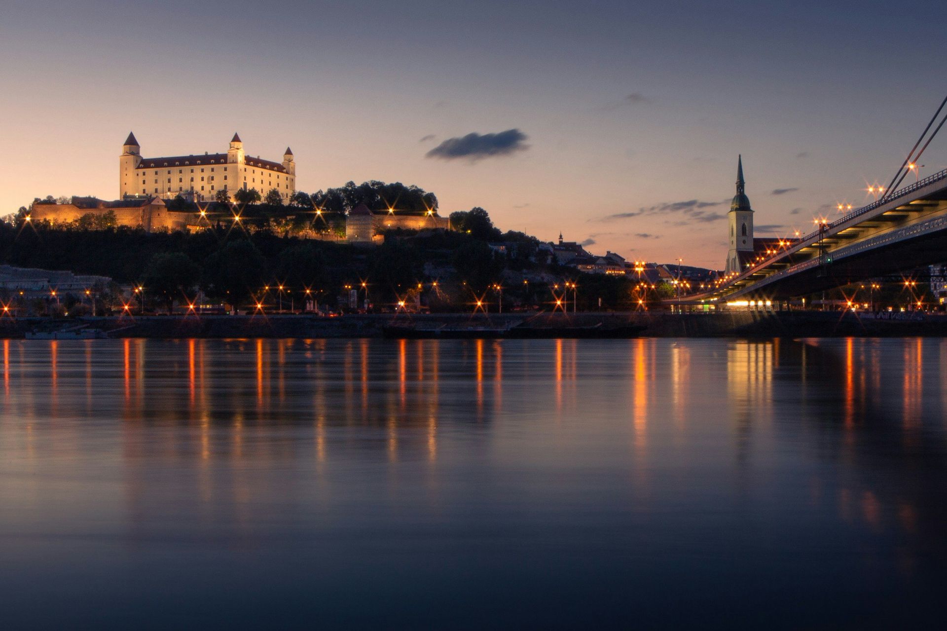Bratislava Castle and cityscape at dusk, lights reflecting on the Danube River.