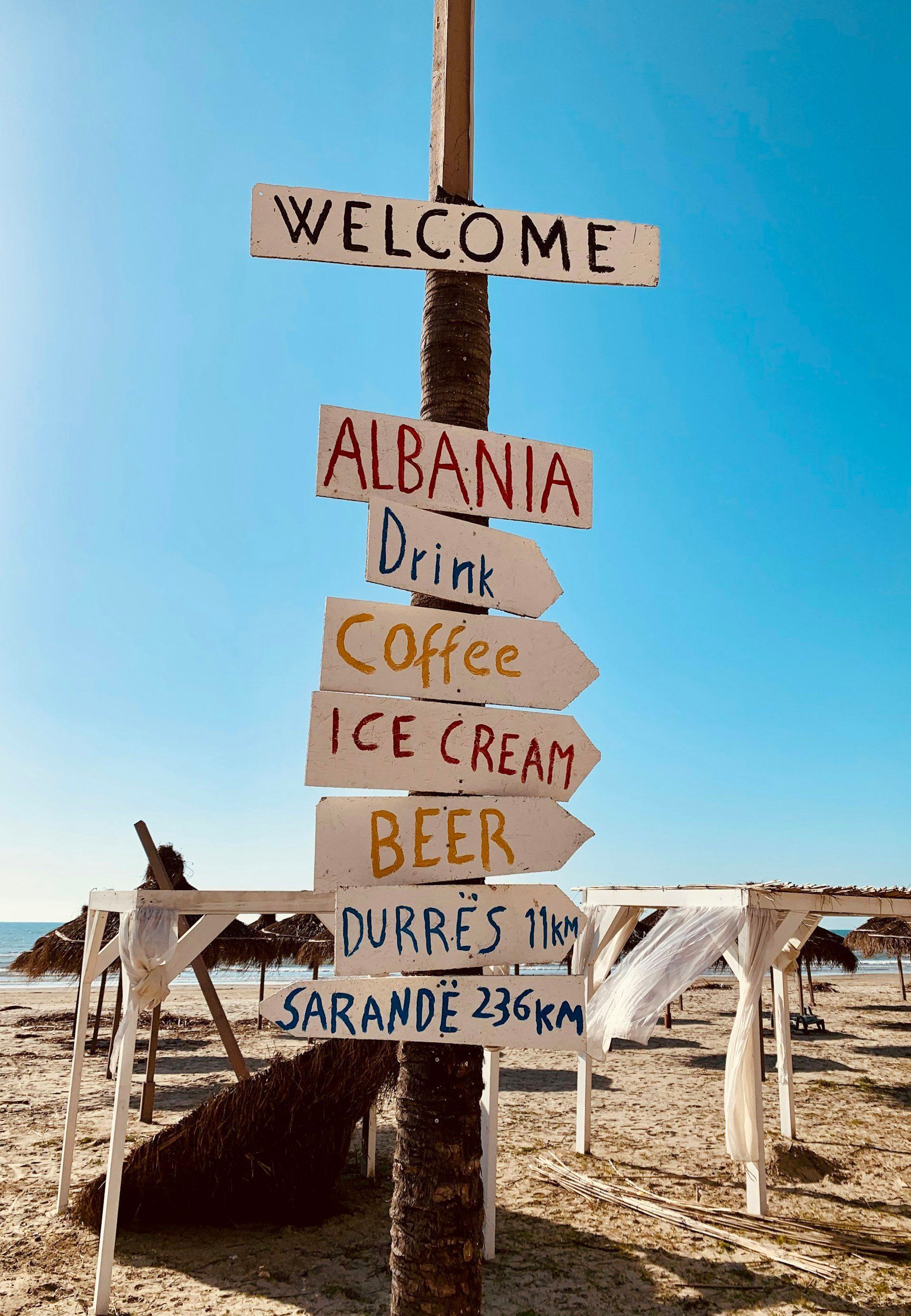 Signpost on a sunny beach, pointing to Albania, drinks, coffee, ice cream, beer, and nearby cities.