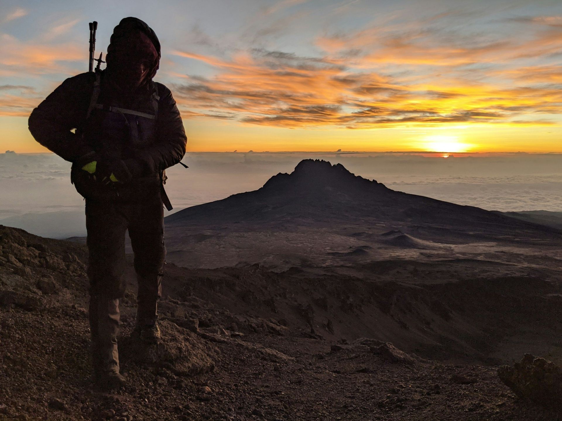 Person on a mountain summit silhouetted against a sunrise. Distant peak and clouds below.