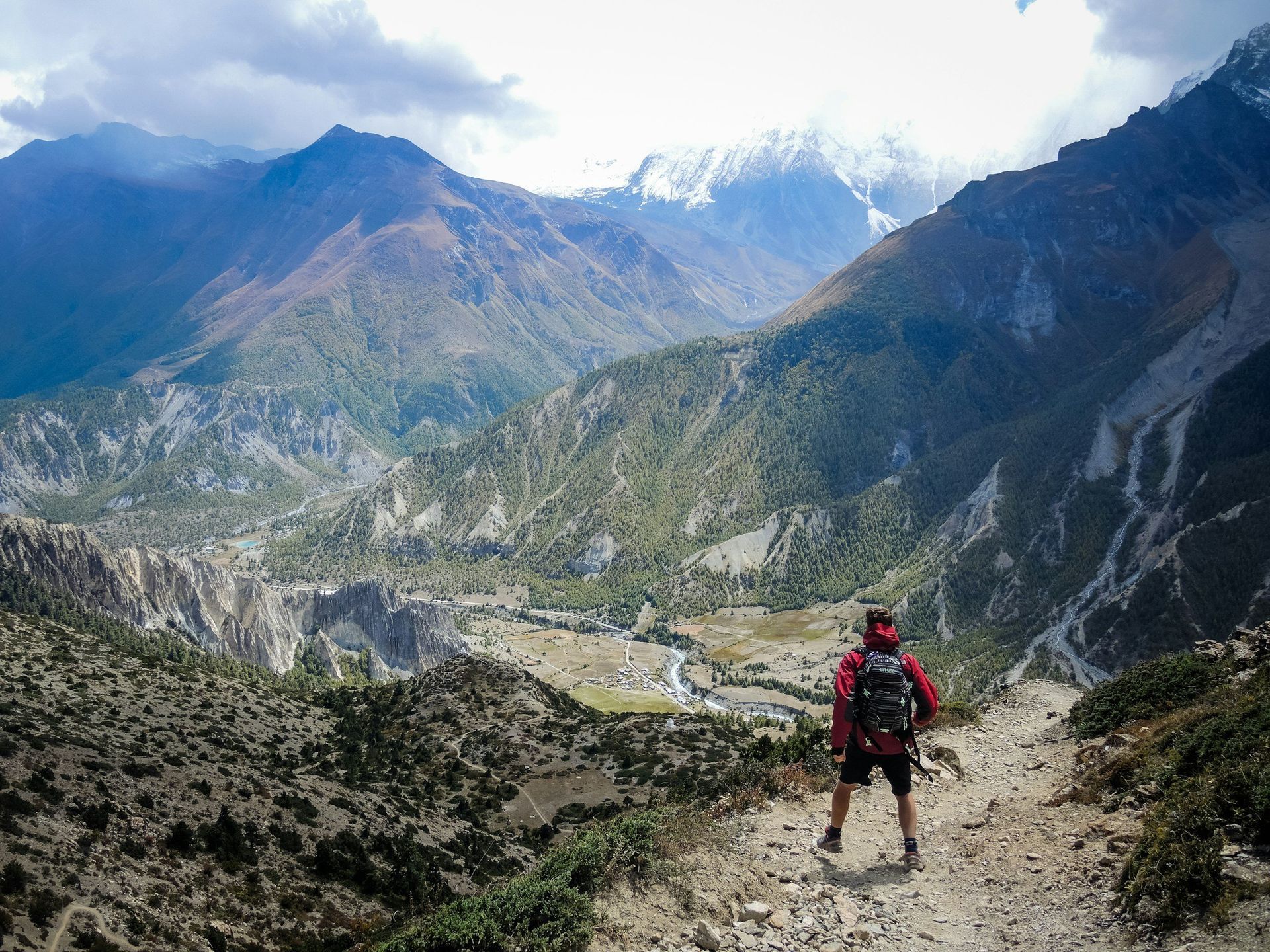 Person hiking on mountain path, overlooking valley with village and snow-capped peaks.