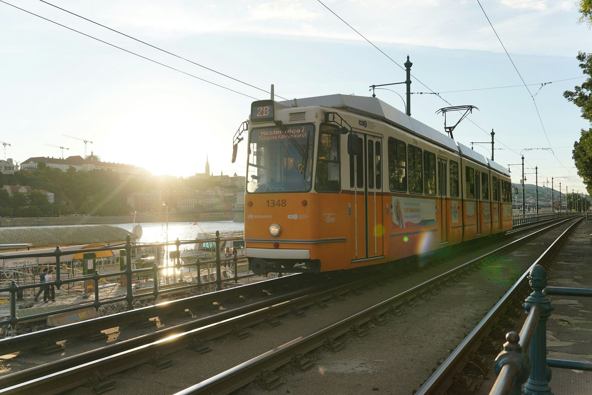 Orange tram on tracks, sunlit, beside a river; city and skyline in the background.
