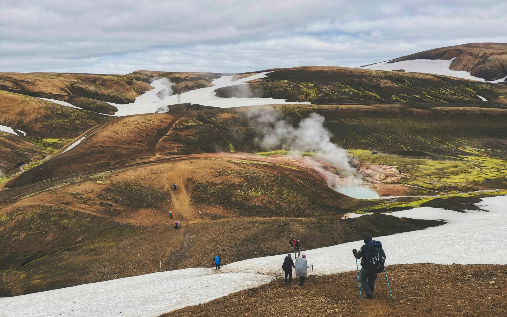 Hikers traverse a snowy, mountainous landscape with steam rising from geothermal vents.