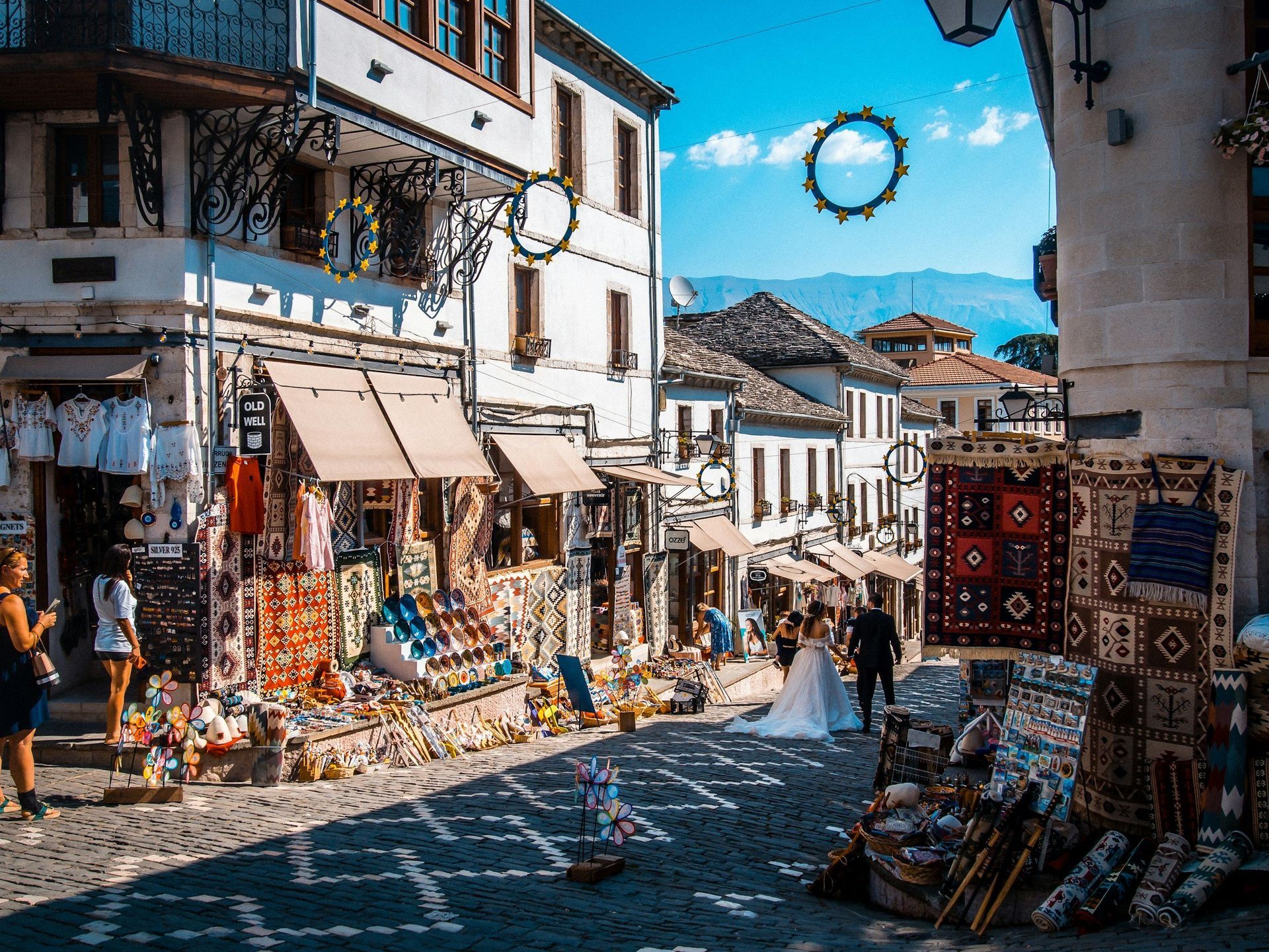 Cobblestone street lined with shops, two people in wedding attire, sunny day, mountains in the distance.