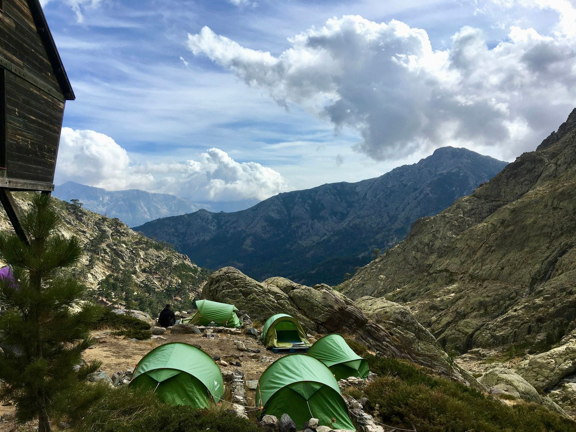 Green tents set up on a hillside overlooking a mountain range under a cloudy sky.