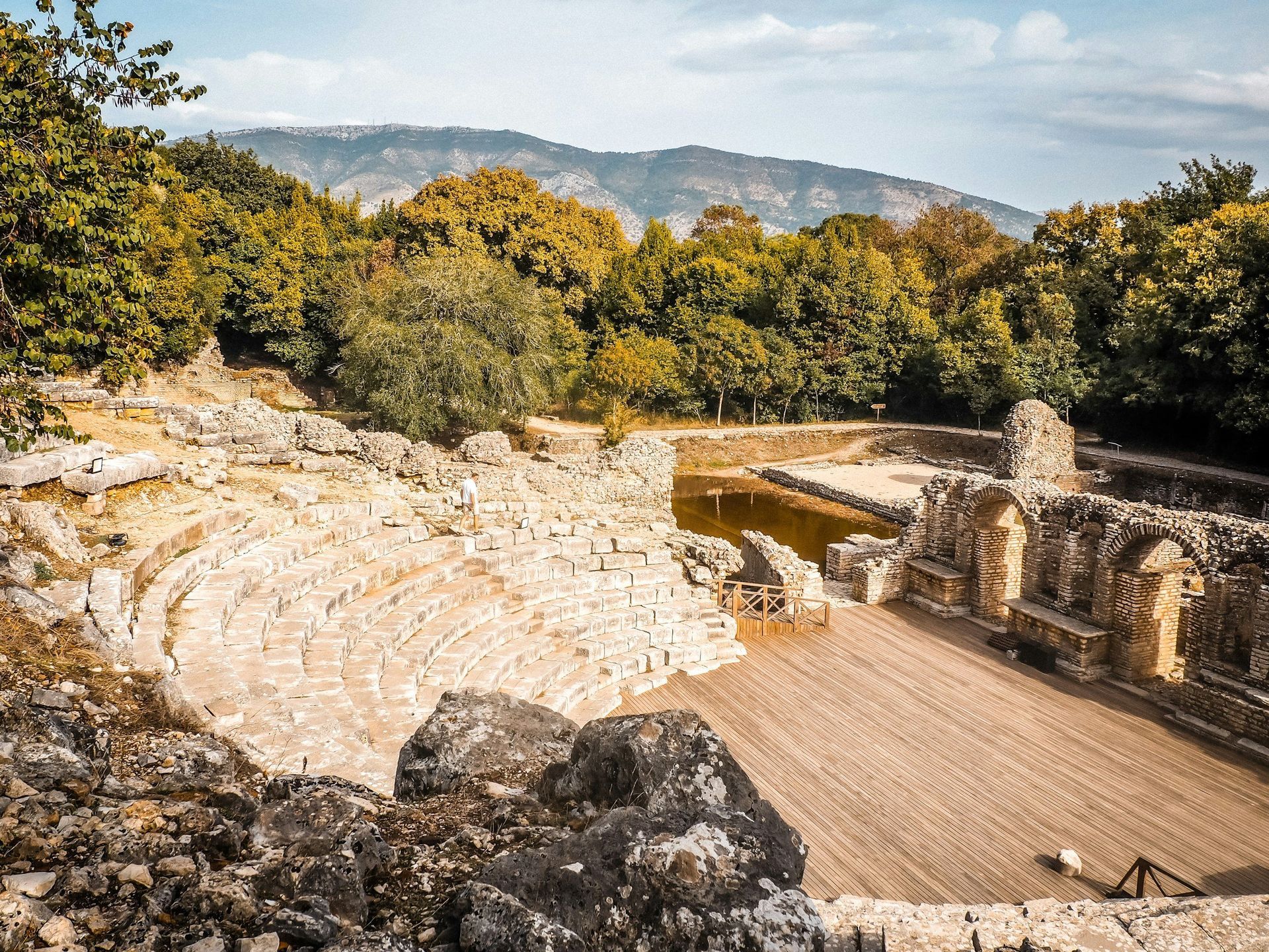 Ruins of an ancient amphitheater with stone seating, facing a stage area. Trees and mountains in the background.