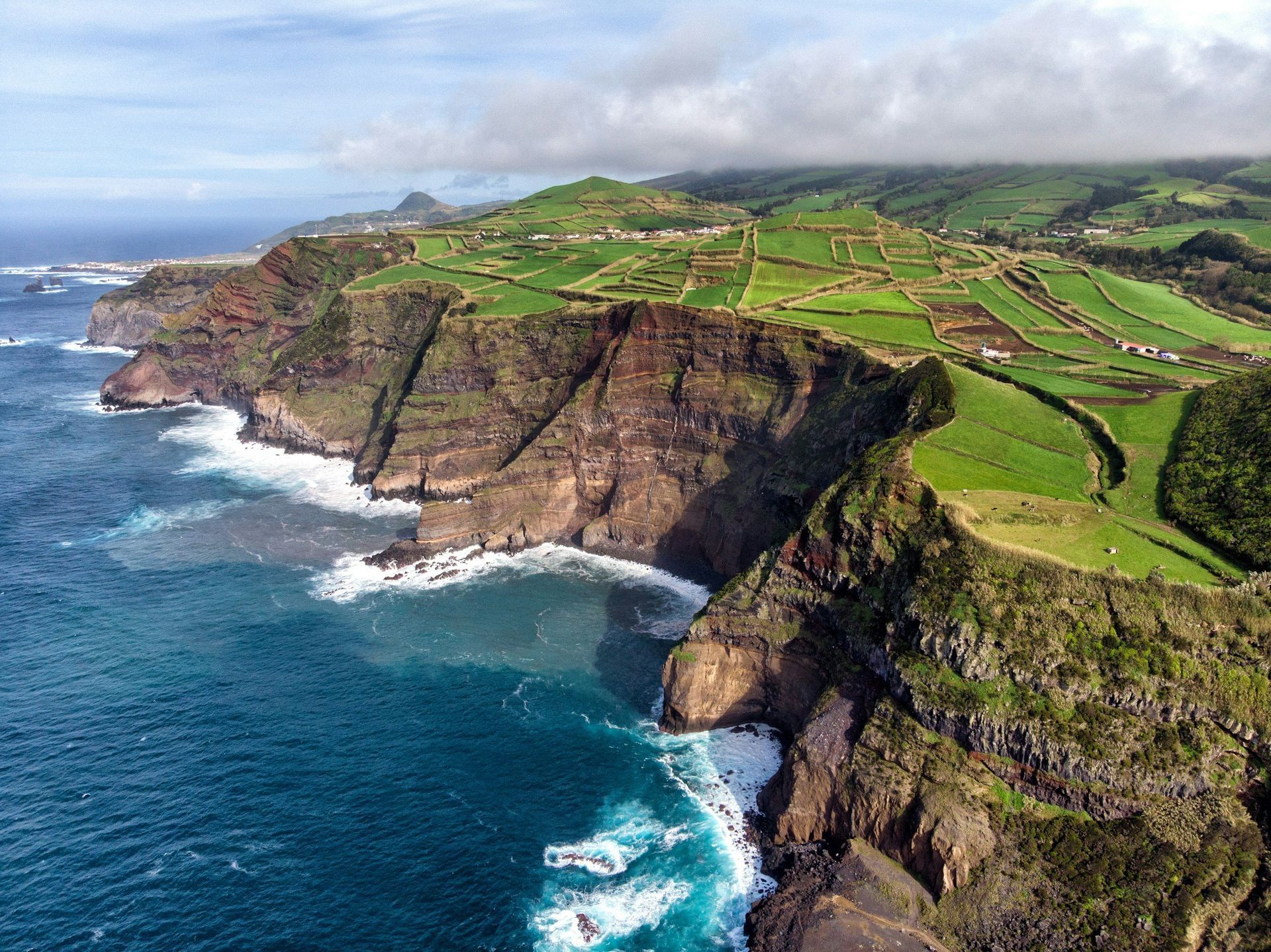 Rugged coastline with cliffs and green fields meeting the blue ocean under a cloudy sky.
