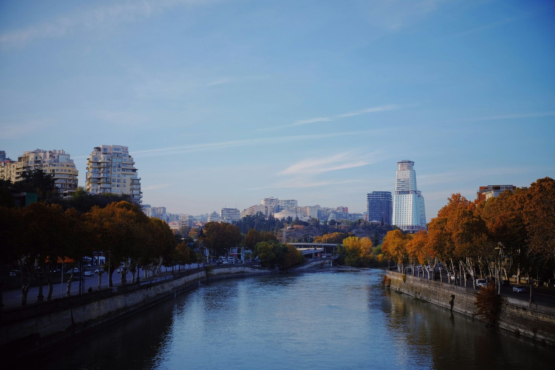 Cityscape with river, buildings, and autumn trees under a blue sky.