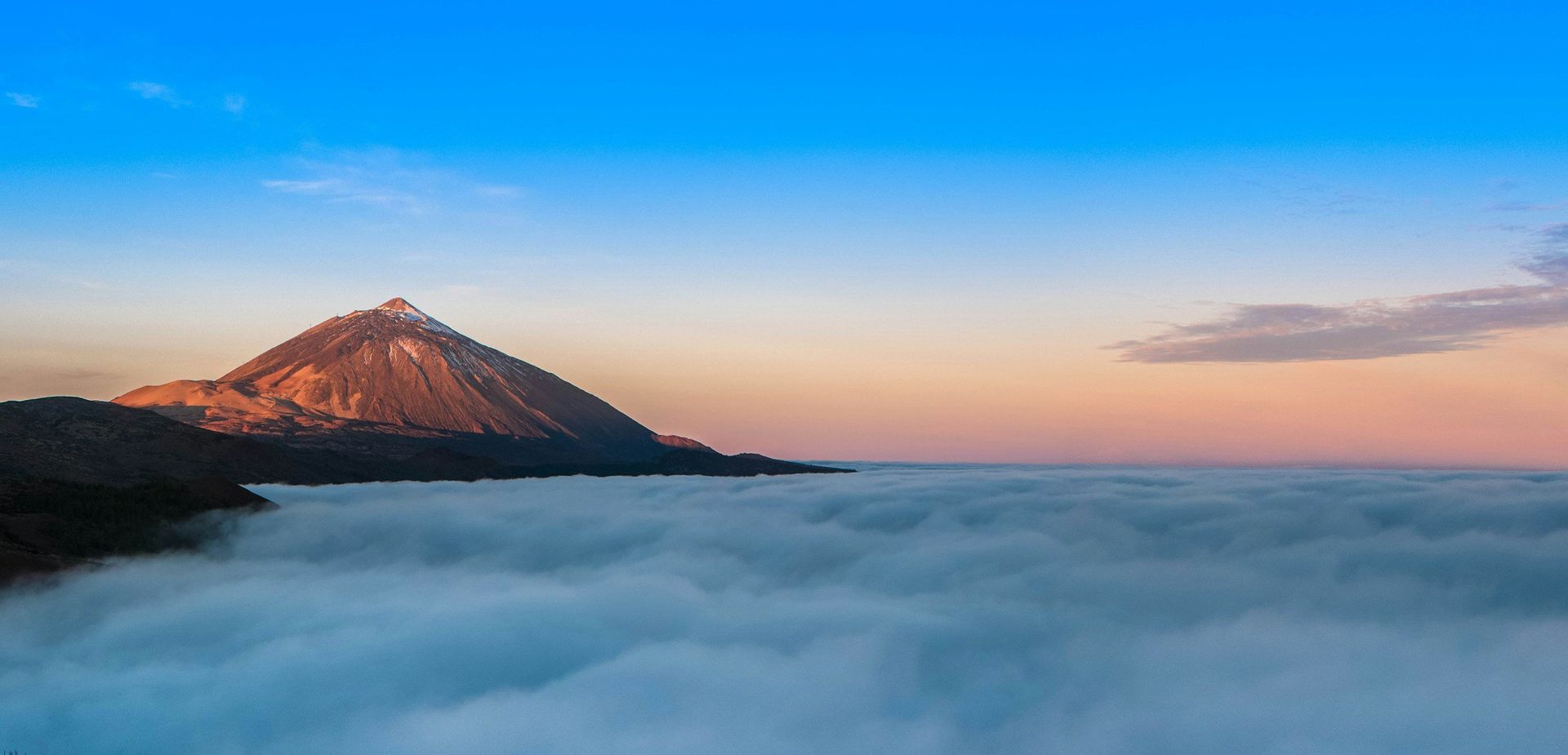 A mountain peak emerges from a sea of clouds under a blue and pink sky.