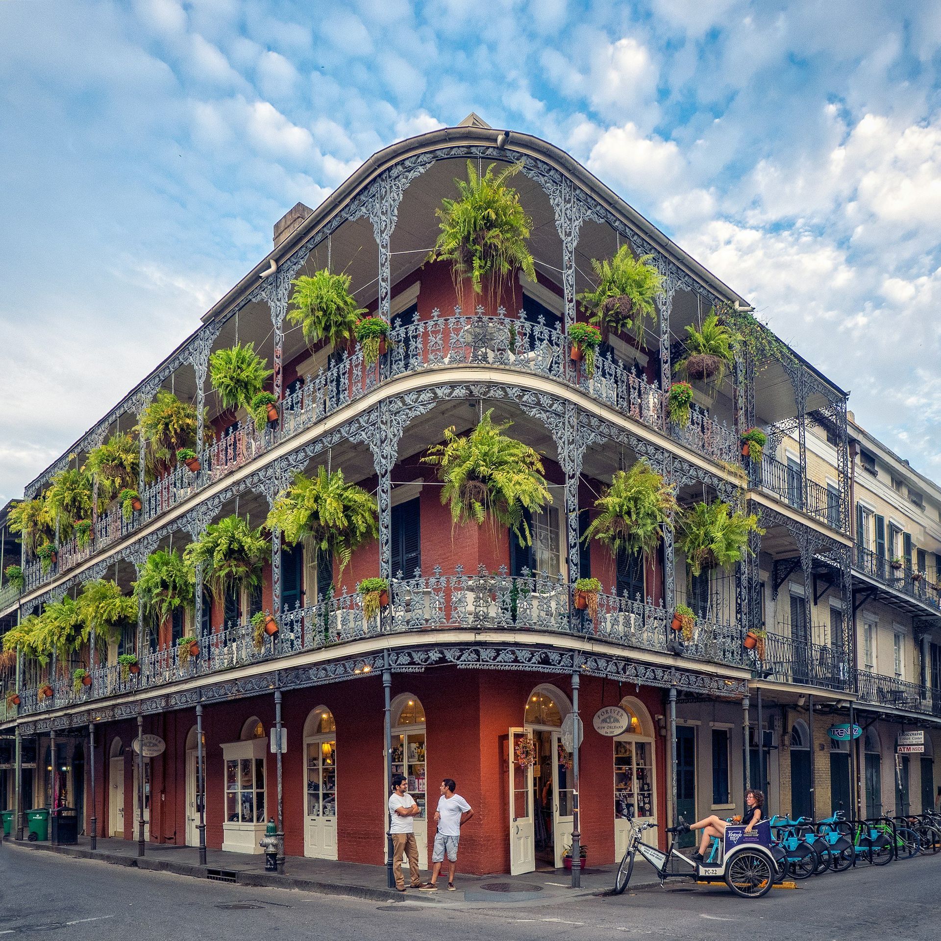 Corner building with ornate wrought-iron balconies covered in plants in New Orleans, with people and bikes.
