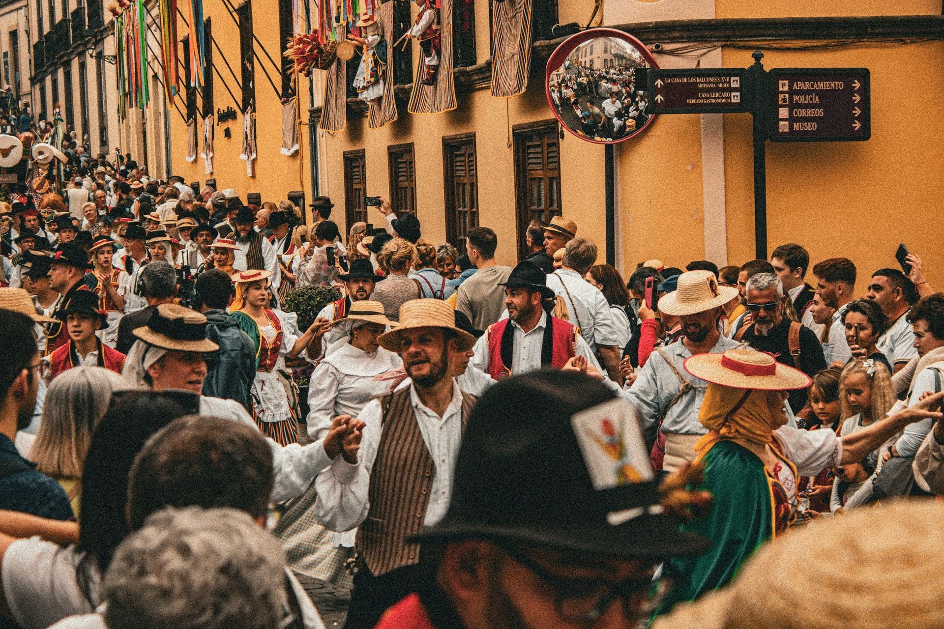 Crowd celebrating in a narrow street, buildings with flags, many wearing hats, lively atmosphere.