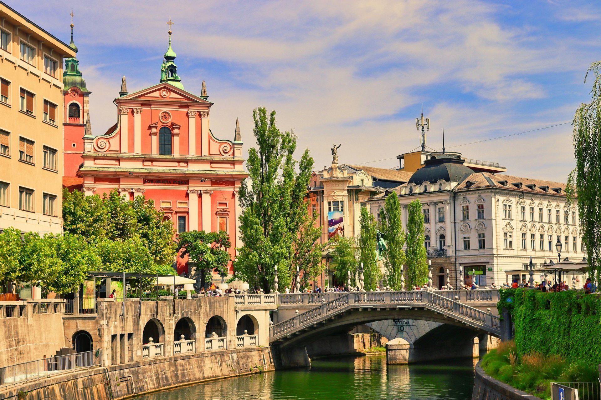 View of Ljubljana, Slovenia, with a bridge, river, and buildings, including a pink church, on a sunny day.