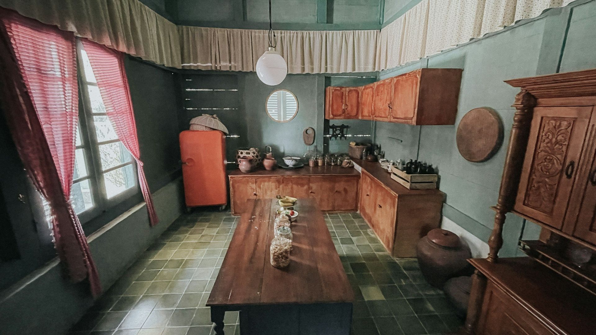 Rustic kitchen with wood cabinets and table, orange refrigerator, red curtains, and green tiled floor.