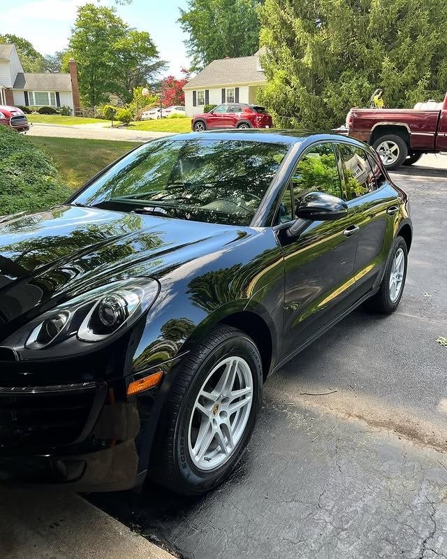 Black Porsche Macan SUV parked on a driveway with a red pickup truck and houses in the background.