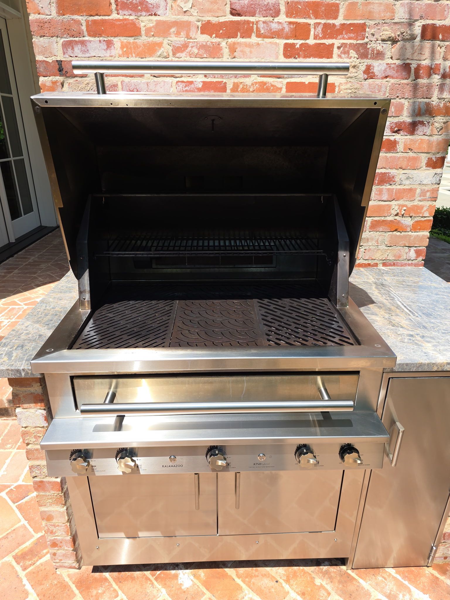 Stainless steel outdoor grill with open lid, set against a brick wall.