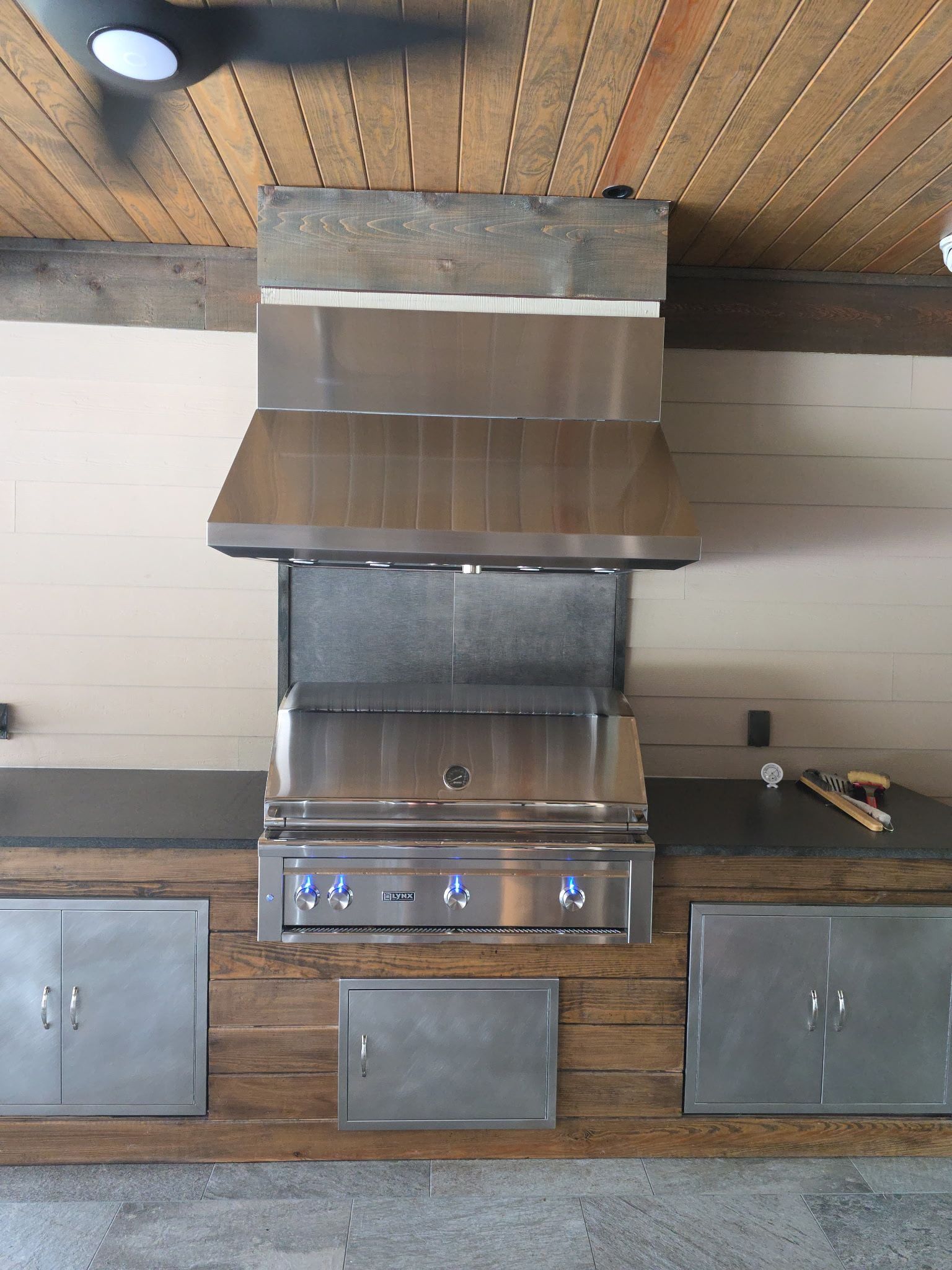 Outdoor kitchen with a stainless steel grill, hood, and storage cabinets built into a wooden and stone structure.